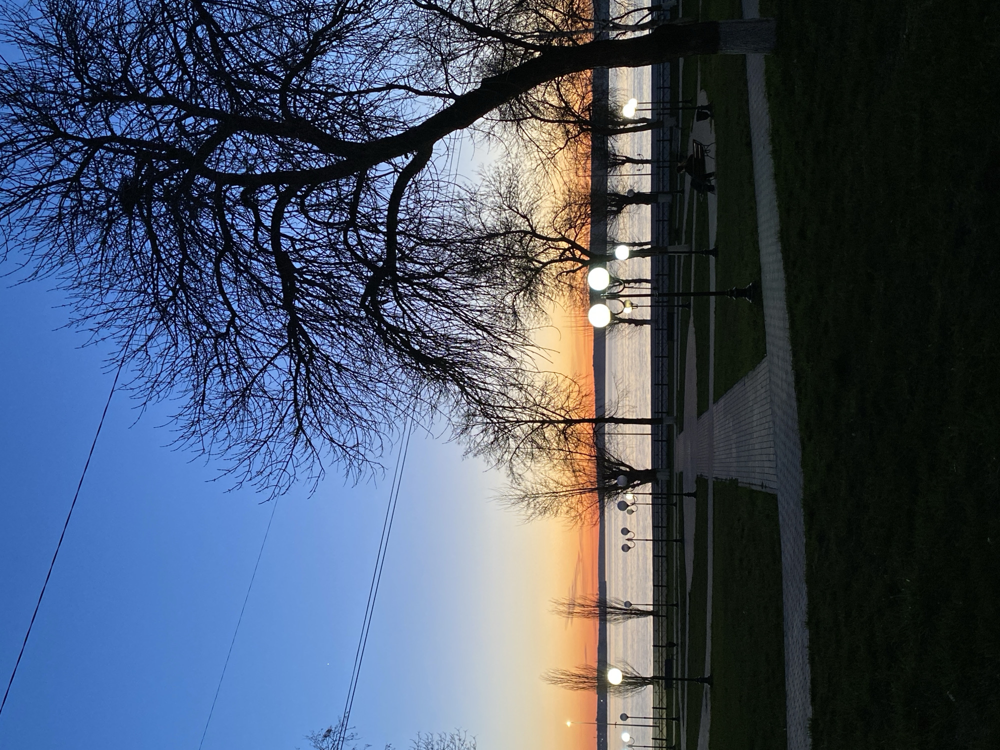 The image depicts a waterfront scene in Votkinsk, Russia, at twilight or dusk. A row of spherical streetlights illuminates a paved path alongside what appears to be a body of water, possibly the Votkinsk Reservoir. Bare trees frame the scene, their branches silhouetted against the sky. The sky exhibits a gradient from blue to orange and yellow, indicative of sunset or sunrise. The path is bordered by green grass and possibly park features. The scene is generally still and tranquil, without apparent human activity in the foreground. Overhead power lines are visible to the lower left of the image.