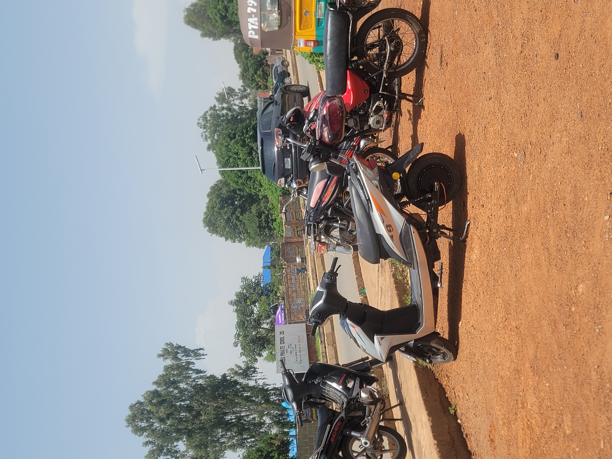The image depicts an outdoor scene in Barakin Lerawi, Nigeria, on a sunny day. Several motorcycles and a scooter are parked on an unpaved, reddish-brown surface in the foreground, with a curb to the left. Behind them, a street is visible with a dark SUV, a sedan, and a three-wheeled vehicle (likely an auto-rickshaw) parked on the right side. In the background, trees and buildings are present, including a sign for "SACRED PRIVATE SCHOOL JOS." The sky is clear and blue. No people are prominently featured or engaged in any specific activity.