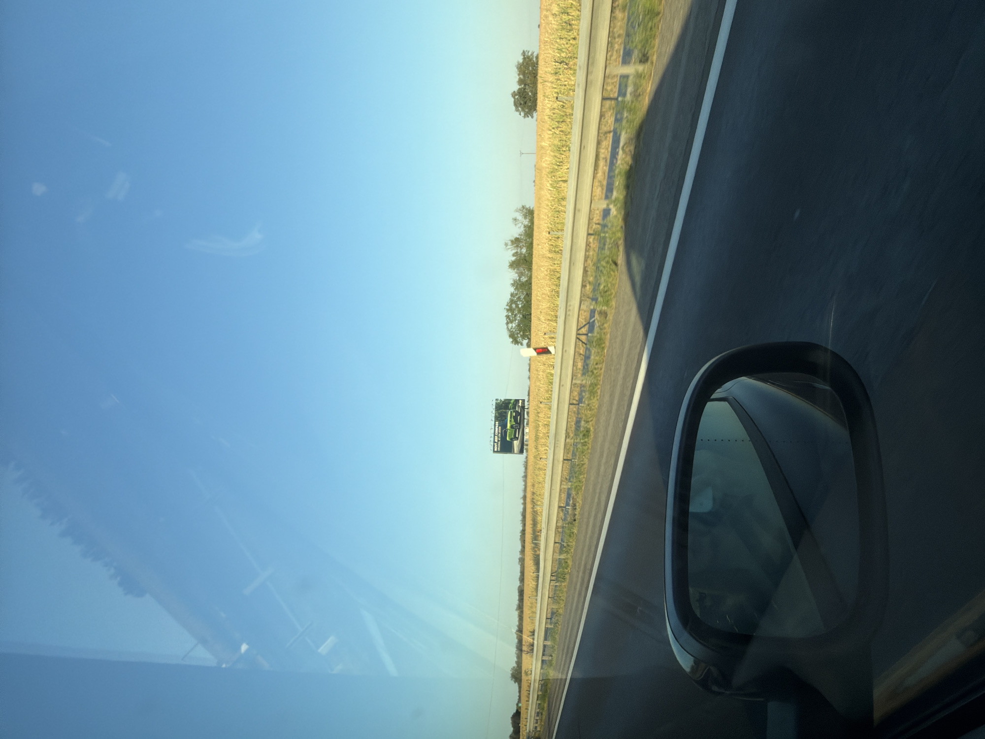 The image captures a roadside view from a vehicle traveling on a highway. The sky is a clear, bright blue, with the sun casting long shadows. On the right side of the highway, a field of dry corn stalks stretches towards a line of trees in the distance. A large billboard advertising a green Jeep Compass is visible in the middle ground, situated above the cornfield. The highway itself has a light grey asphalt surface with a white line marking the lane. A metal guardrail separates the highway from the field. In the foreground, the reflection of the sky and passing scenery is visible on the car's windshield, and the driver's side mirror is also in frame, reflecting a blurry interior. The scene is set in the rural outskirts of Pata, Slovakia, characterized by agricultural land and clear weather.