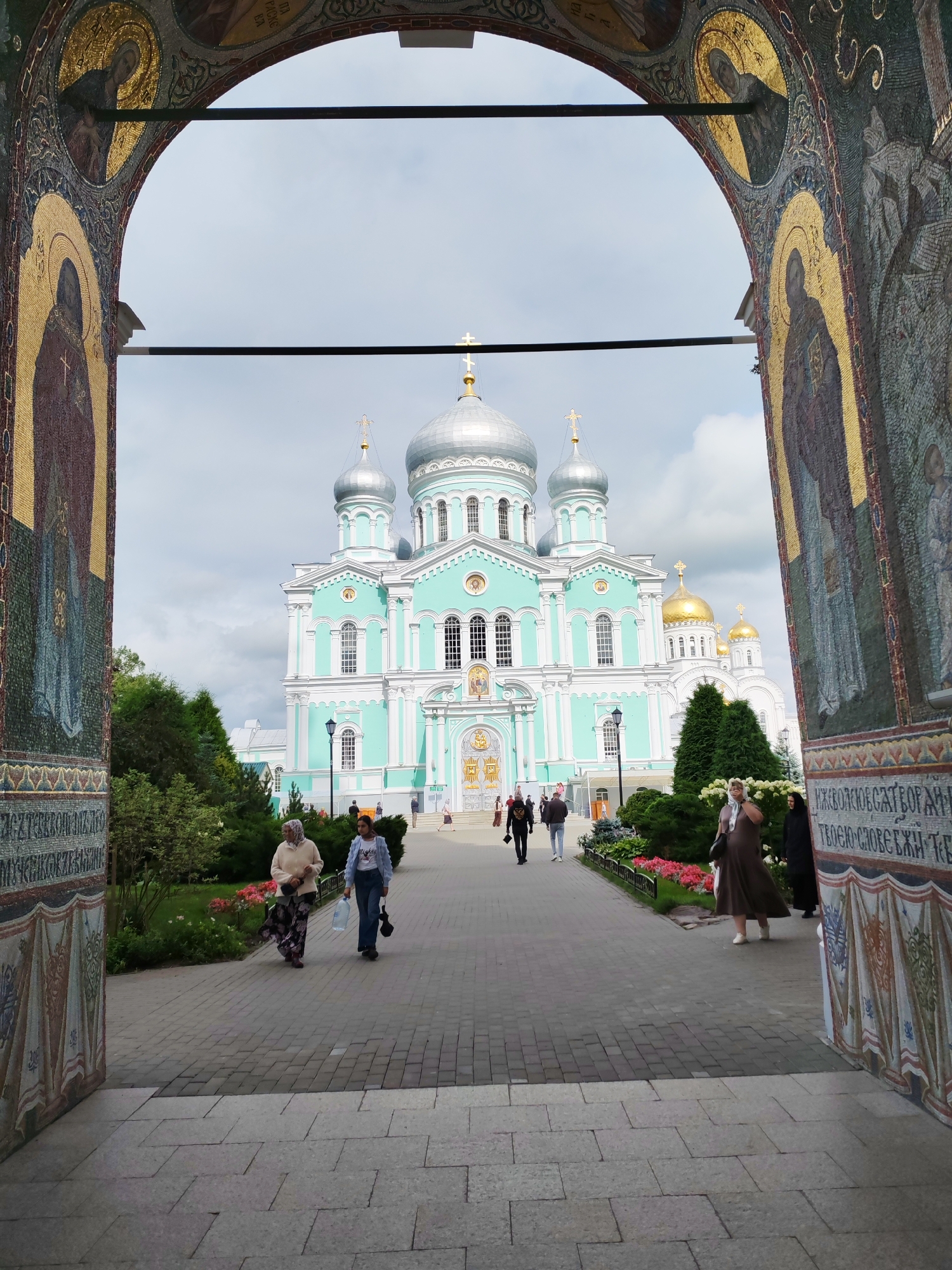 The image depicts the Holy Trinity Cathedral in Diveyevo, Russia, viewed through an ornate archway decorated with mosaics of religious figures. The cathedral, with its distinctive turquoise and white facade and silver domes, stands prominently in the background under a cloudy sky. A paved walkway leads towards the cathedral, bordered by lush greenery and flowerbeds. Several individuals are present, walking along the path and towards the entrance. On the left side of the path, two women are walking together, one in a light-colored headscarf and patterned skirt, the other in a blue jacket and jeans, carrying a plastic water bottle. Further down the path, other figures can be seen, including a person in a black outfit and headscarf. The archway frames the scene, adding a sense of grandeur and religious significance to the view of the cathedral complex.