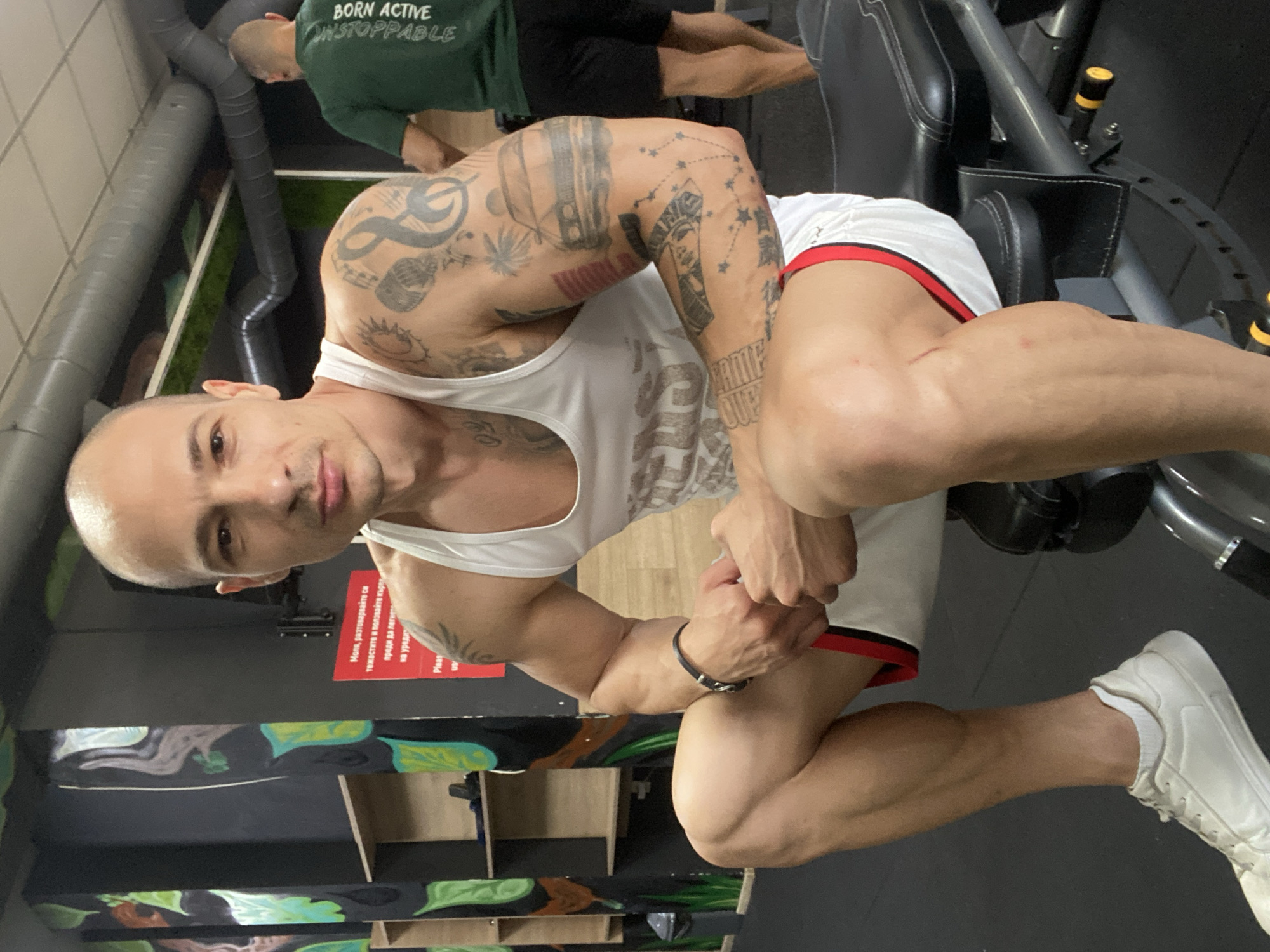 Here is a detailed description of the image:
**Scene:** The photo appears to have been taken indoors, likely in a gym in Pleven, Bulgaria, as indicated by the text in the image.
**People:** The image prominently features a muscular, bald man with multiple tattoos on his arms. He is wearing a white tank top and white shorts with red trim. He is looking at the camera and posing. In the background, there is another man wearing a green shirt with the words "BORN ACTIVE UNSTOPPABLE" printed on the back.
**Subjects/Objects:** Gym equipment is visible in the background, suggesting a fitness facility. The background also includes wall art with green, blue, and brown leaf-like designs, shelving units, and a red sign with text in Bulgarian. The man in the foreground is wearing a wristband.
**Actions/Interactions:** The man in the foreground is posing for the photo, flexing his muscles. The man in the background is possibly exercising, although the details of his activity are obscured.
**Environment:** The gym setting is evident through the presence of the exercise equipment. The room seems to have a modern design with the wall art and shelving. The lighting is bright.