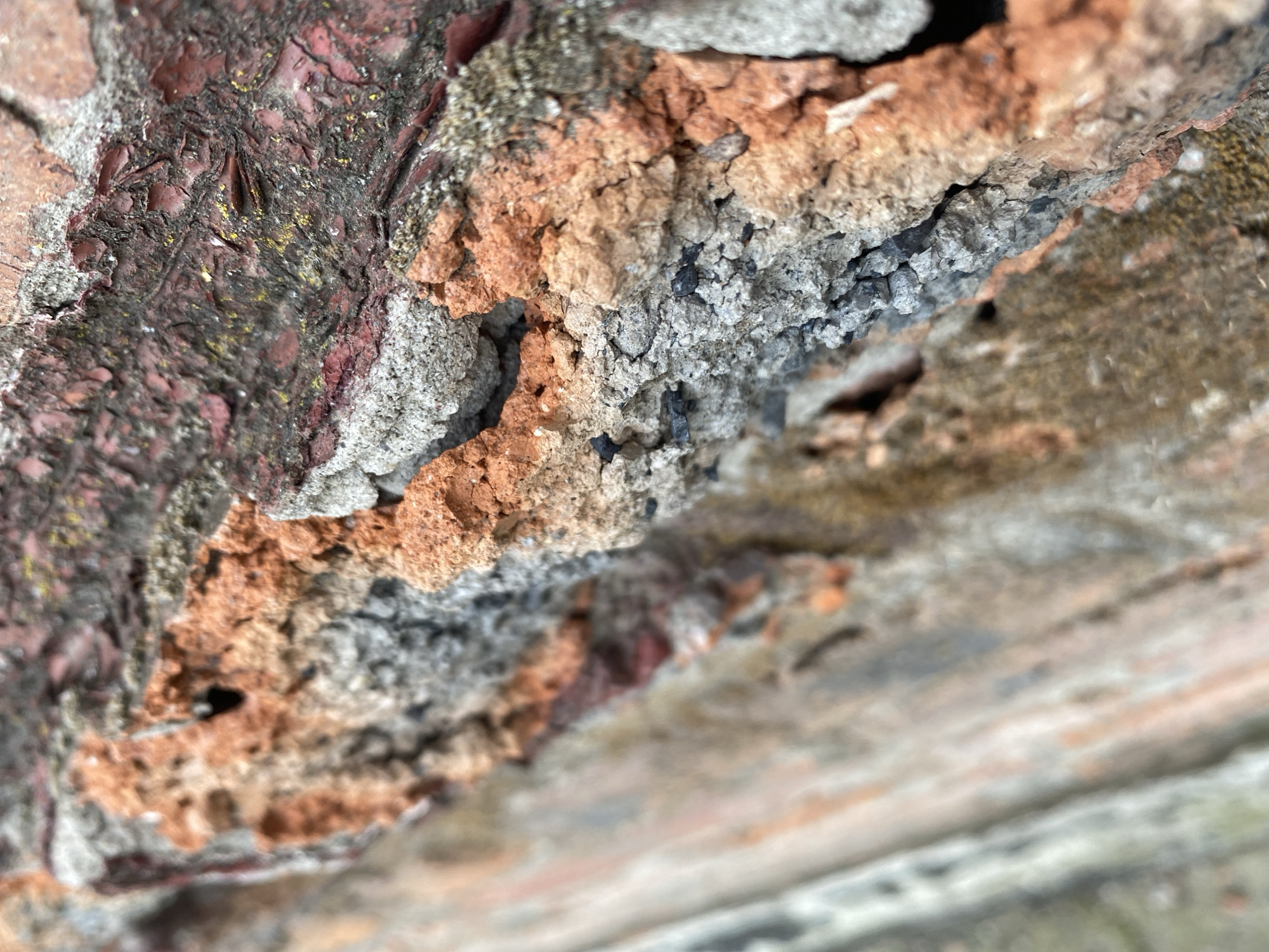 The image is a close-up, macro shot of a weathered brick wall, showcasing its textured surface. The bricks exhibit a range of colors, from deep red to earthy browns and grays, with visible signs of age and degradation. Patches of lighter, granular material, likely mortar or a similar binding agent, are interspersed among the bricks, some appearing crumbly and eroded. There are also small, dark inclusions, possibly fragments of stone or debris, embedded within the material. The lighting emphasizes the rough, uneven surfaces and creates shadows that highlight the depth and porosity of the brickwork. The overall impression is one of age and decay. The image does not depict any people, actions, or interactions. It is a static scene of architectural deterioration. The image is not located in Kisliakovskaya, Russia.