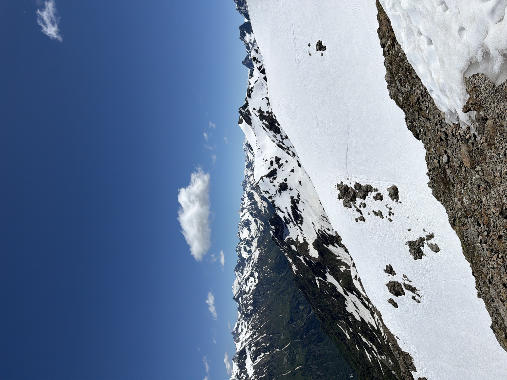 The media file depicts a vast mountain landscape under a clear blue sky. The primary subject is the rugged mountain range, characterized by steep slopes and jagged peaks. Patches of snow are scattered across the mountains, contrasting with the dark rock and green vegetation on the lower slopes. The scene suggests an outdoor environment, likely a natural park or remote wilderness area, given the lack of man-made structures.
The composition of the image implies a sense of scale and grandeur, with the mountains stretching into the distance. The bright sunlight indicates it is daytime, and the clear sky suggests fair weather. The presence of snow implies a high altitude or a season with cold temperatures, likely summer in a high mountain region. The location context provided, "Верхне-Тебердинское сельское поселение, Russia," points to the area being in the Karachay-Cherkess Republic of Russia, known for its mountainous terrain and natural beauty.
There are no visible people or objects in the image, and no discernible activity or event is taking place. The focus is solely on the natural landscape. Notable details include the sharp contrast between the snow-covered and rocky parts of the mountains, the distinct layering of the terrain from green slopes to snow-capped peaks, and the small, scattered clouds in the expansive blue sky. The overall impression is one of serene isolation and the imposing beauty of nature. No text is visible in the media file.