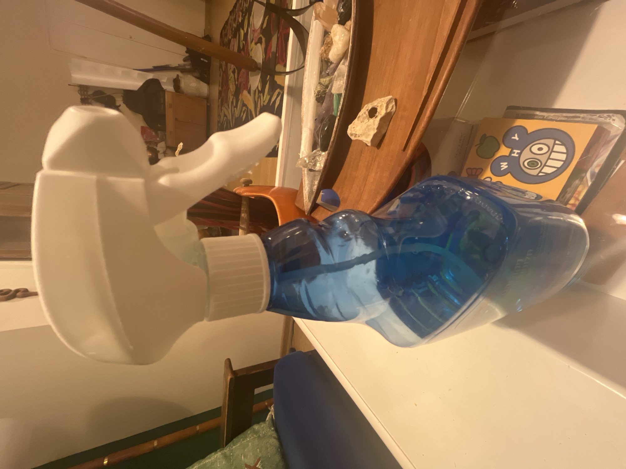 A close-up shot of a blue spray bottle with a white nozzle is on a white surface. A blue cushion and a green patterned pillow are visible below the bottle. In the background, there is a wooden chair, a cabinet, a wooden plank, and various decorative items. There is no evidence that supports this image was taken in Tel-Aviv, Israel.