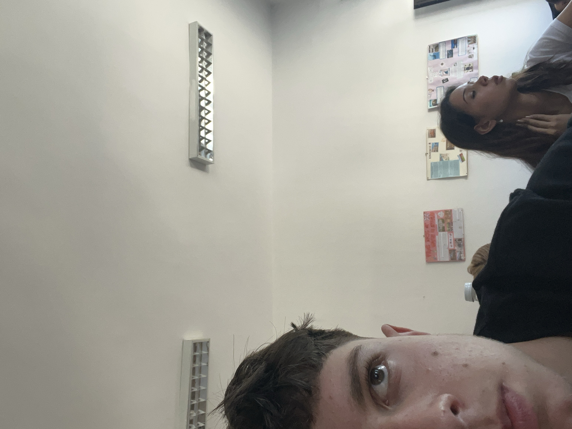 The image shows a close-up of a young person's face and hair in the foreground, with another person and posters on the wall in the background. The young person's left eye and part of their nose and mouth are visible. They have short, dark, messy hair. In the background, a young woman with long, dark hair is looking away from the camera. She is wearing a white shirt and appears to be wearing an earring. Behind them, on the wall, are several posters with text and images. The environment appears to be indoors, possibly a classroom or common area, with fluorescent lighting fixtures visible on the ceiling. There is no indication of the location being in Zlatograd, Bulgaria.