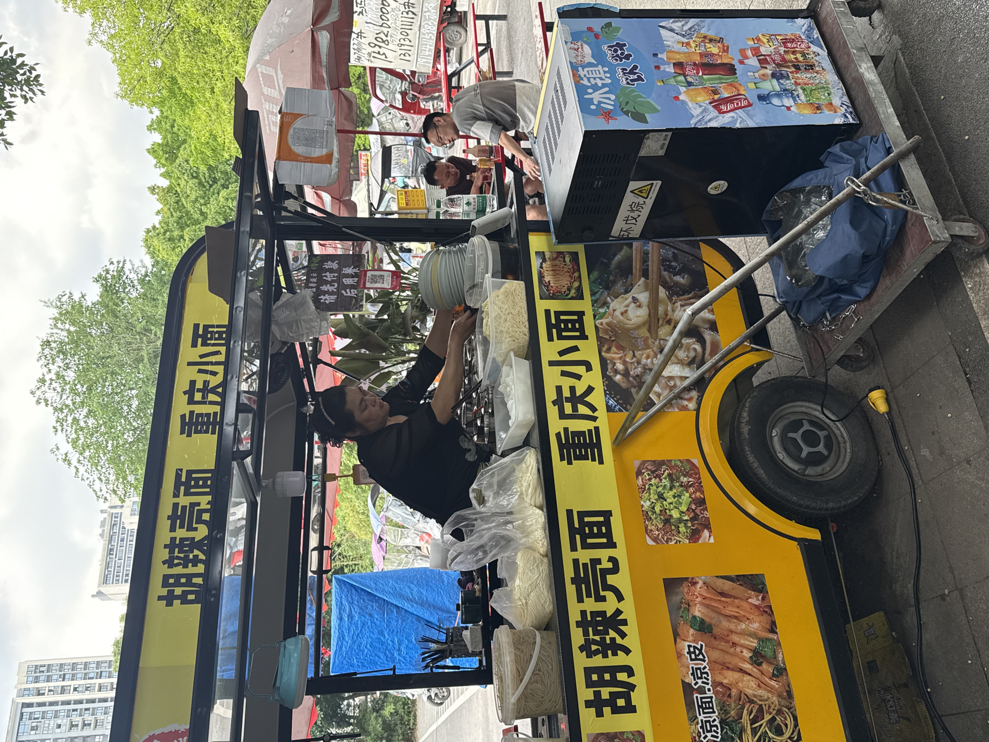 A food vendor is at a street-side stall in Chongqing High-tech Zone, China. A woman is serving food from the mobile cart that has "胡辣壳面 重庆小面" on the side, which translates to "Hu spicy shell noodle Chongqing small noodle". The cart also has images of different types of noodles, including "凉面.凉皮", which translates to "Cold noodles. Cold skin". A cooler sits next to the cart, filled with bottled beverages, and the sign says "冰镇饮料", which translates to "Iced Drinks". The vendor is serving two men in the background. An electrical cord is coming from the cart. Signs in the background state to pay first before eating.