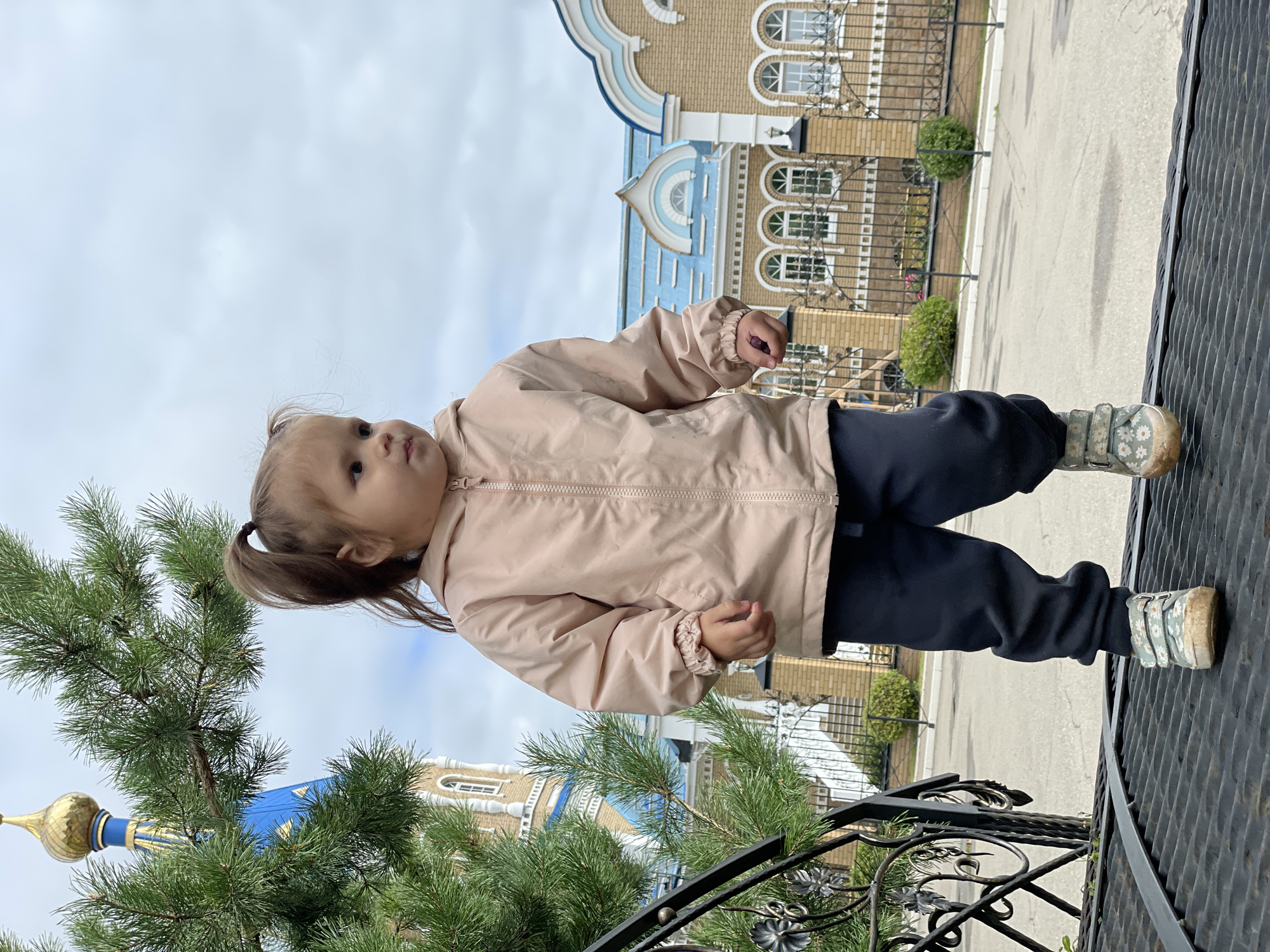 A young girl with a brown ponytail stands on a black metal grate in front of a building with a blue roof and a golden dome, likely a church or religious building. She is wearing a light pink jacket, dark pants, and floral-patterned sneakers. A pine tree branch partially obstructs the view of the building. The sky is cloudy. This scene appears to be located in Izhevsk, Russia, given the architectural style of the building.