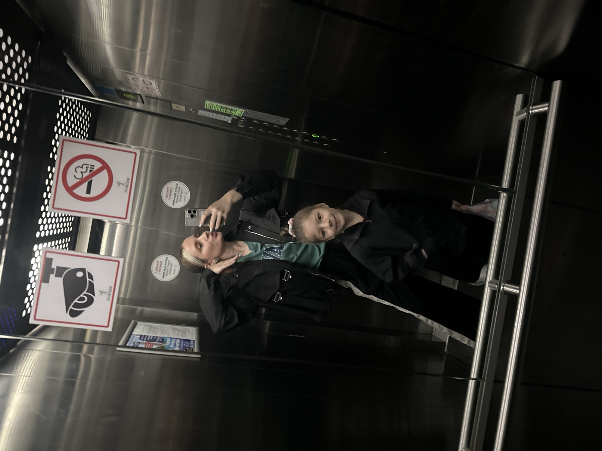 Here is a detailed description of the image:
**Scene Overview:**
The image captures a selfie taken inside an elevator. The elevator appears to be in Esenyurt, Turkey, as indicated by the Turkish text and "Akkóza" branding visible on the signage within the elevator. The interior of the elevator has a metallic finish, with safety signs posted on the walls. The lighting in the elevator is somewhat dim.
**People:**
Two individuals are present in the image. The older one appears to be a young adult, possibly in their late teens or early twenties. The younger one is a child, seemingly around 6-8 years old. The woman is holding a phone and taking a selfie while holding the little girl.
**Objects and Details:**
- Elevator features: No smoking sign, safety signage with stylized illustrations, a panel displaying elevator information, and vertical metal bars along the side.
- Personal items: The older individual is wearing a black jacket with a black purse or small bag. She also wears a light green shirt.
- Clothing: Dark-colored clothing worn by the people in the image, with a white line on it.
**Actions and Interactions:**
The primary action is the taking of a selfie in the elevator. The woman holds her phone in one hand while posing with the child.
**Environment:**
The environment is contained within the elevator. The metal surfaces, signage, and safety features suggest a modern building or complex within Esenyurt, Turkey.
