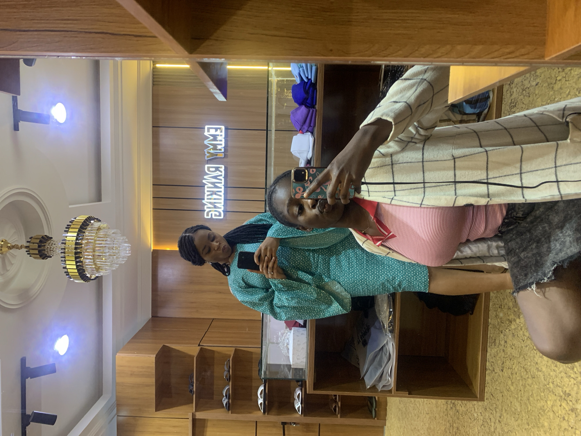 In a well-lit boutique or clothing store in Gura Top, Nigeria, two young women are captured in a mirror selfie. The store features warm wooden shelving units filled with merchandise, including shoes and folded apparel. A prominent neon sign spelling "EMMY RANKING" in a distinctive font is mounted on the back wall.
The woman in the foreground, positioned closer to the mirror, is kneeling and holding a smartphone up to take the picture. She is wearing a pink and white striped collared shirt under a cream-colored, checkered open cardigan. Her dark hair is pulled back, and she is looking directly into the camera, her lips pursed.
Standing behind her, in the reflection, is another woman wearing a vibrant teal-green dress with a subtle white polka dot pattern. The dress has wide, flowing sleeves. She is holding a smartphone in her right hand, also seemingly taking a picture, and is looking down at her phone with a focused expression.
Above them, a decorative chandelier with gold accents and crystal elements hangs from the ceiling. Track lighting is also visible, contributing to the bright illumination of the store. The flooring appears to be a textured yellow material. The overall atmosphere suggests a modern retail environment.