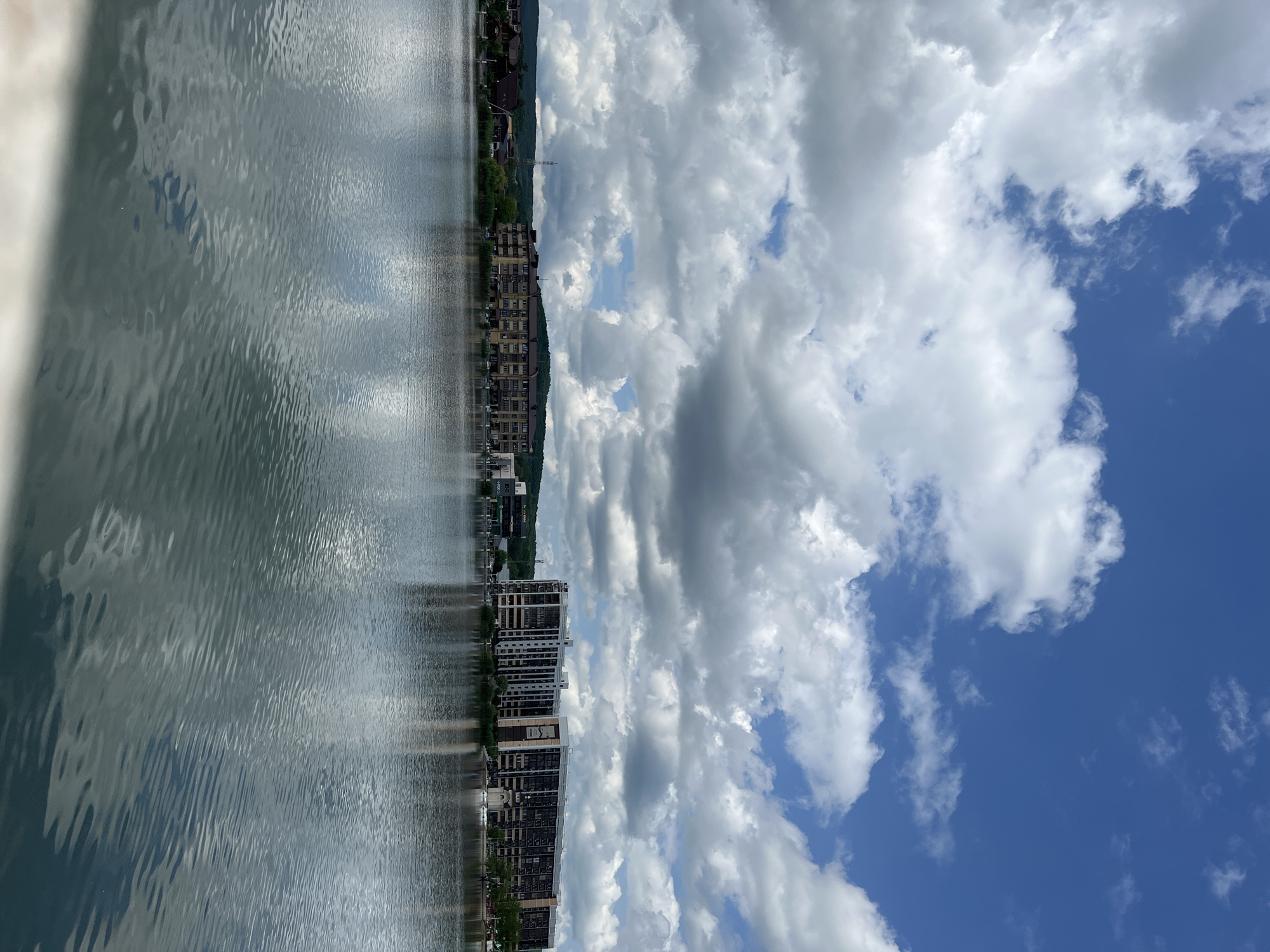 The image depicts a body of water reflecting buildings, trees, and a cloudy sky. The location appears to be a waterfront area in the city of Goryachiy Klyuch, Russia. Buildings of various heights are lined up along the waterfront, possibly residential or commercial. The water shows reflections of the structures and the sky above. The sky is filled with puffy white clouds against a blue backdrop. There is no human activity visible in the image. The environment seems to be a mix of urban development and natural elements.