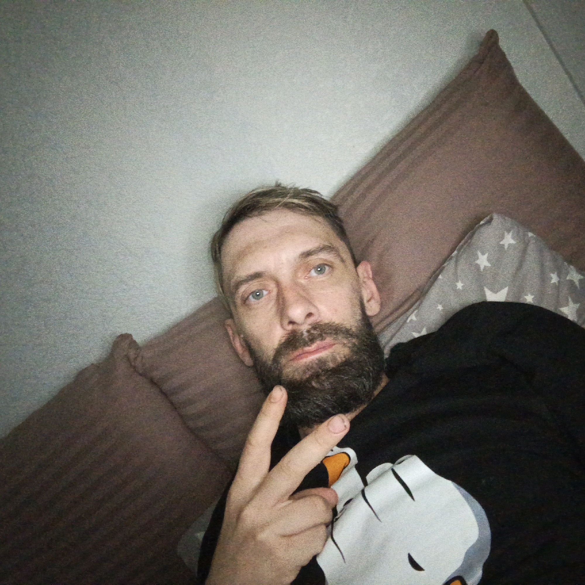 A man with a full dark beard and light-colored hair lies in an indoor setting, likely a bedroom or living room, in Markovskiy, Russia. He appears to be taking a selfie, holding up his right hand in a peace sign towards the camera. He wears a dark hoodie or shirt featuring a white and orange graphic, which resembles a stylized penguin character. His expression is neutral to slightly contemplative.
Behind him are pillows; one is a solid brown or mauve color, and another is gray with a pattern of white stars. The background consists of a plain, light-colored wall. The lighting in the image is somewhat dim, giving the photograph a slightly grainy quality, suggesting it may have been taken in low-light conditions, possibly in the evening or at night. Notably, his fingernails are visible, appearing unmanicured. No discernible text is present in the image.