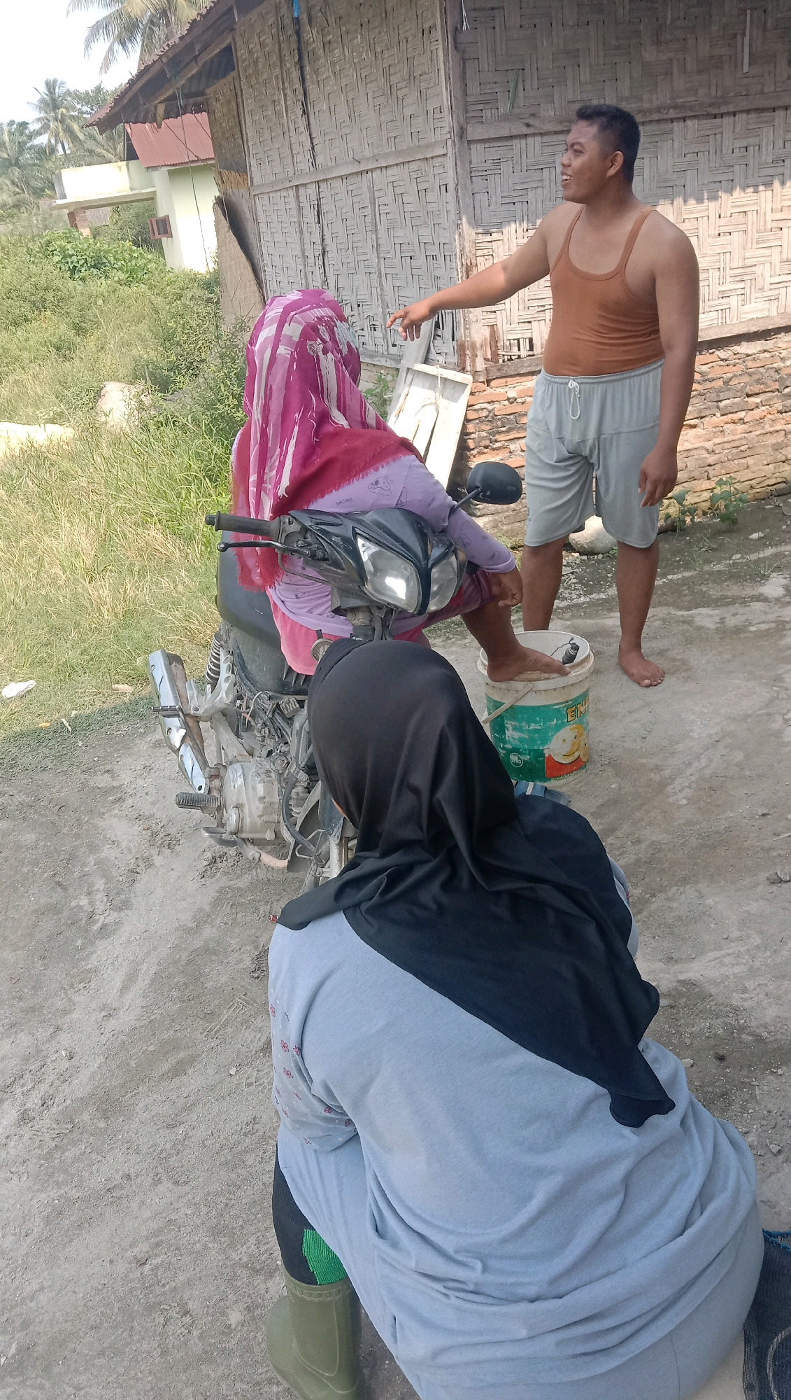 In a village setting that appears to be in or near Kota Pari, Indonesia, three individuals are interacting near a motorcycle and a rustic wooden structure. One person, a male wearing a brown tank top and grey shorts, is standing and gesturing towards a person seated on a motorcycle. This seated person is covered by a pink and maroon hijab and appears to be wearing a purple top. A third individual, facing away from the camera and wearing a black hijab and a grey long-sleeved shirt, is kneeling near the motorcycle. The environment is characterized by dirt ground, overgrown grass, and palm trees visible in the background, suggesting a rural or semi-rural location. A green bucket with some labeling is present near the standing man, and one of his feet is placed on it. The motorcycle is parked on the dirt ground, and its components, such as the exhaust and engine, are visible. The overall scene depicts a casual interaction outdoors.