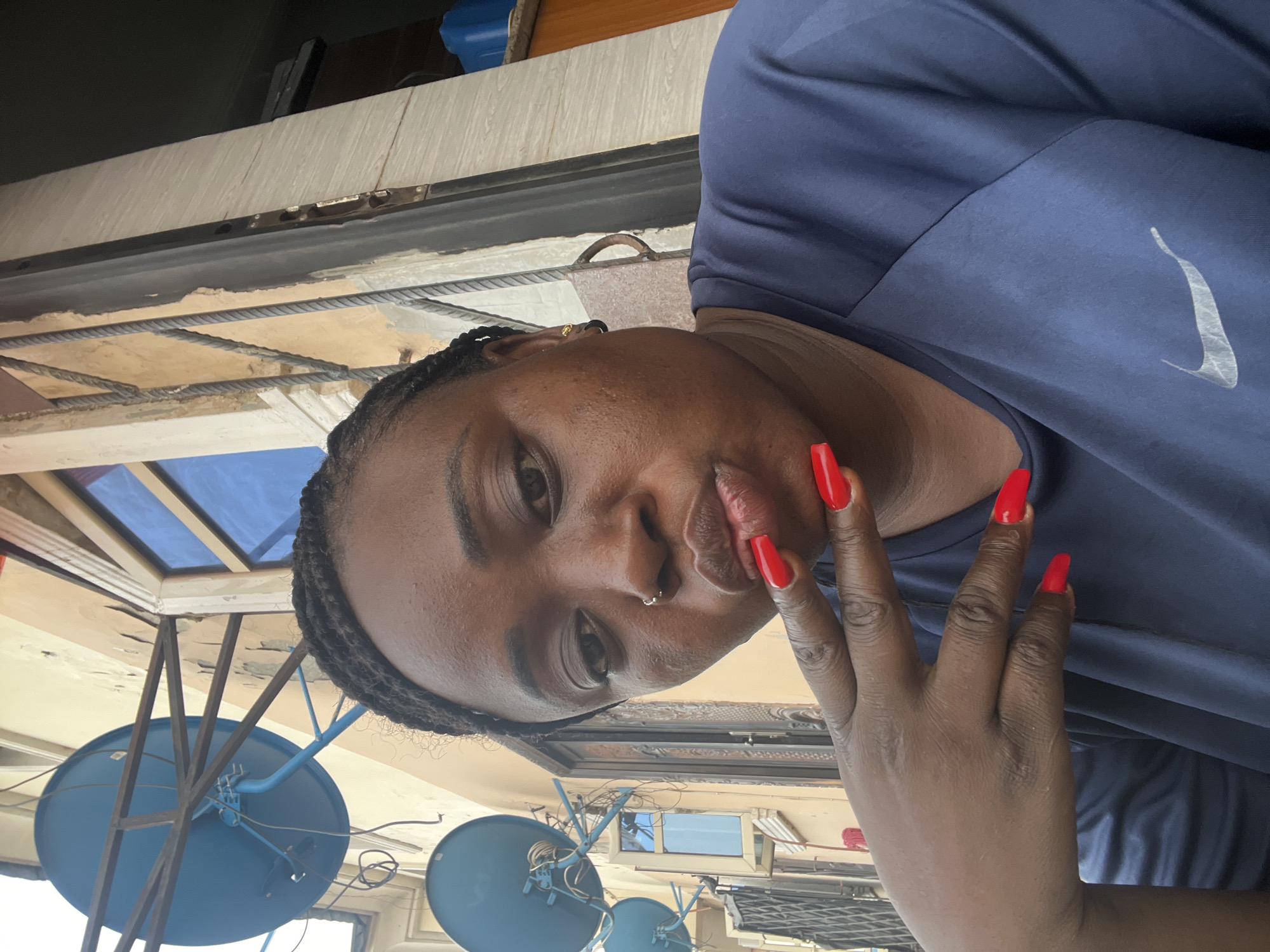 A medium close-up angled shot shows a person with dark skin and braided hair. They are wearing a dark blue t-shirt featuring a Nike swoosh logo on the upper right. Their nails are long and painted red. They have a nose ring on their left nostril. The person is looking at the camera, and their hand is near their face.
In the background, there is an overhang with a glass roof supported by metal beams. Also visible are multiple blue satellite dishes mounted on metal frames. The location appears to be an outdoor setting, possibly a rooftop in Dushepe, Nigeria.
