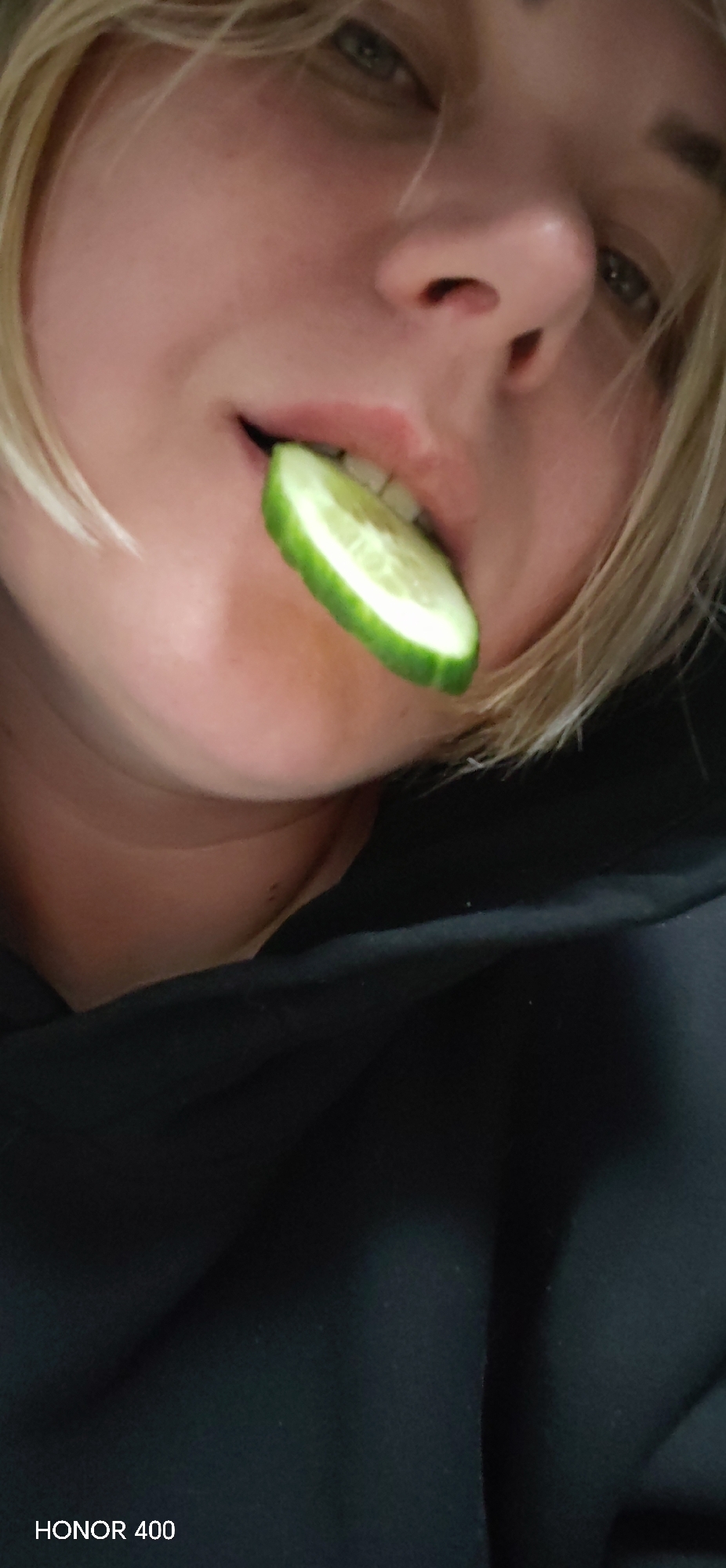 A person with light skin and blonde hair is holding a slice of cucumber in their mouth. The camera angle is close-up and looking upwards at the person's face, capturing details such as their eyes, nose, and the cucumber slice between their lips. The person is wearing a black garment, likely a hoodie or sweater, which occupies a large portion of the lower part of the frame. The background is dark and undefined. The text "HONOR 400" is visible in the bottom left corner, indicating the device used to take the photo. There is no information to suggest the photo was taken in Noviy, Russia.