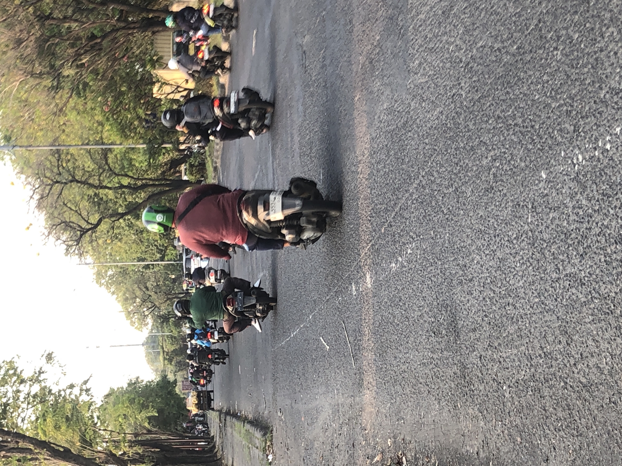 The image depicts a street scene with numerous motorcycles and riders in Gelam, Indonesia. The perspective is from behind a rider on a motorcycle, looking forward down a paved road lined with trees. The road is partially obscured by traffic, consisting primarily of motorcycles. Several riders are visible, many wearing helmets. The environment appears to be a moderately busy urban or suburban area, likely during daytime, given the natural light. The road surface is asphalt and shows signs of wear. The trees lining the road suggest a green, possibly well-maintained, area.