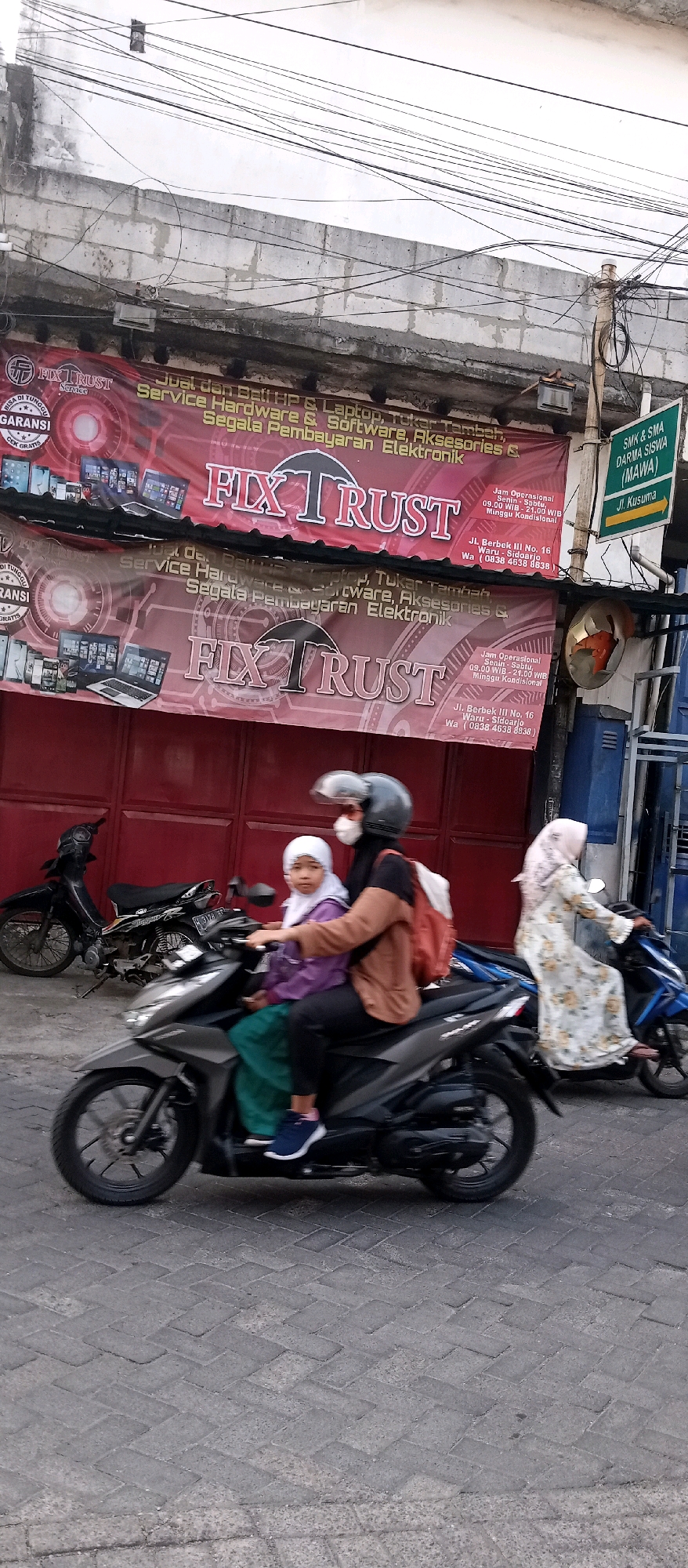 In Wadungasri, Indonesia, a street scene unfolds with two motorbikes prominently featured. The foreground displays a dark-colored motorbike carrying two individuals: an adult driver wearing a helmet, mask, and a brown jacket over a black shirt, and a child passenger in the front, also wearing a helmet and a purple jacket over a white headscarf and green pants. Behind them, another motorbike, a black one, is parked.
Above the street, a weathered concrete wall is visible, adorned with numerous electrical wires creating a dense, tangled network. Two large red banners with white and pink lettering are hung below the wall. The top banner reads "FIX TRUST" and advertises services such as "Jual dan Beli HP & Laptop, Tukar Tambah, Service Hardware & Software, Aksesoris & Segala Pembayaran Elektronik." The bottom banner is a duplicate of the top one.
To the right of the banners, a green street sign with white lettering points to "Jl. Kusuma" and indicates "SMK & SMA DARMA SISWA (MAWA)." Further to the right, a circular chrome mirror reflects the scene. The ground is paved with interlocking grey bricks laid in a herringbone pattern. The lighting suggests it is daytime, possibly late afternoon, with soft, diffused light.