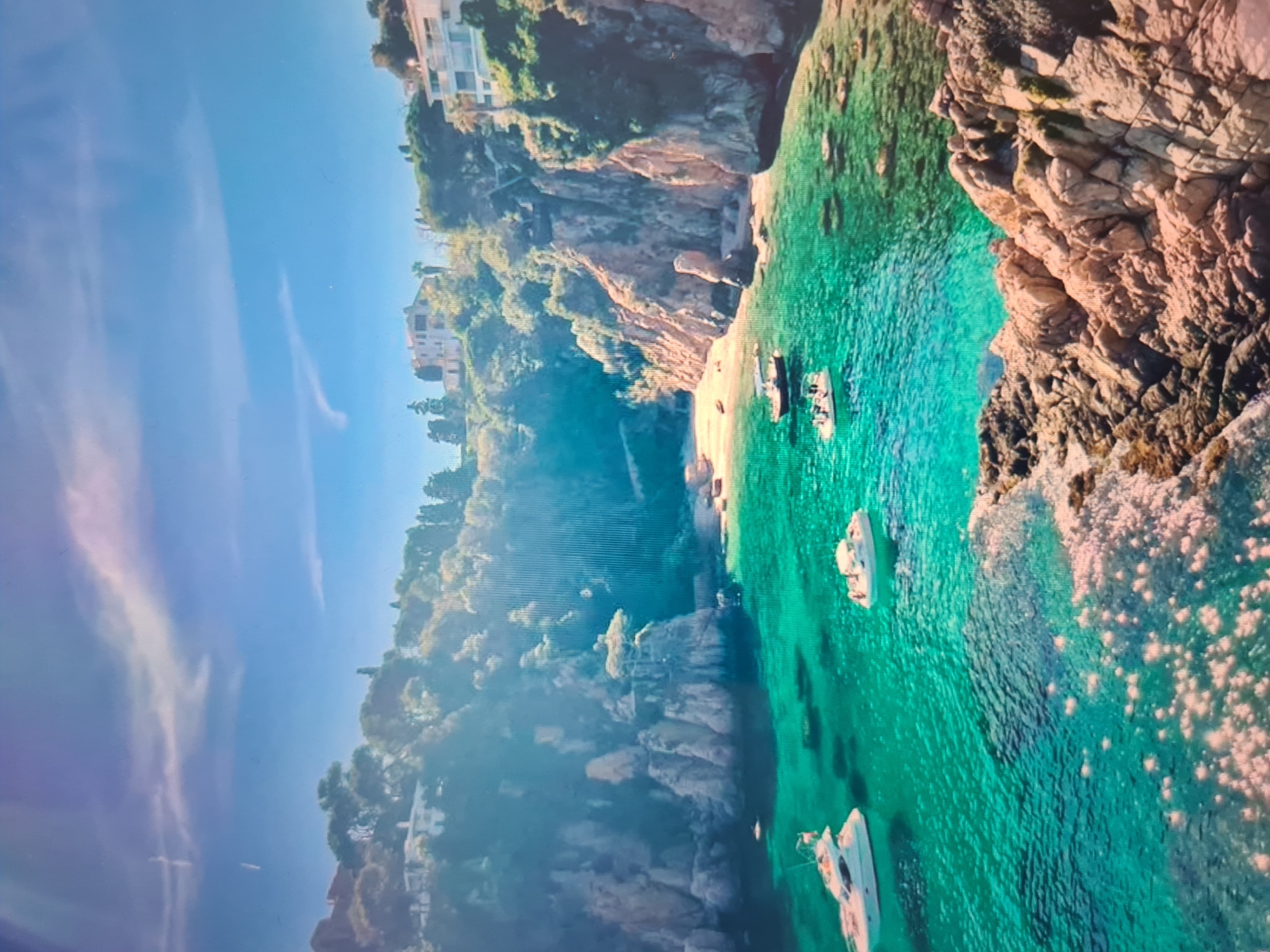 This image presents a stunning aerial or high-angle view of a serene coastal landscape, bathed in bright daylight. The scene showcases a picturesque cove nestled against steep, rocky cliffs.
In the upper portion of the frame, a clear, vibrant blue sky is visible, adorned with a few wispy, stretched-out white clouds. Below the sky, the rugged coastline is dominated by imposing cliffs densely covered in lush green Mediterranean vegetation and trees. Perched atop and along these cliffs are several elegant white buildings, likely villas or houses, with flat roofs and balconies, offering panoramic views of the sea. Some man-made structures and pathways are faintly discernible descending the cliff face towards the water.
The coastline forms several secluded, sandy coves at the base of the cliffs. The water is remarkably clear and a brilliant turquoise-emerald color, allowing the seabed to be seen in shallower areas. Scattered across the calm, transparent water are several small white boats, appearing to be yachts or leisure crafts, anchored peacefully. One larger boat is prominent in the foreground, while others are positioned closer to the sandy shores. Gentle ripples and small areas of white foam from breaking waves are visible where the water meets the rocky shore on the right side.
The overall impression is one of an idyllic, tranquil, and beautiful summer destination. The time of day appears to be sunny late morning or early afternoon, with no people visibly present, emphasizing the untouched beauty and peacefulness of the scene. There is no visible text in the image.