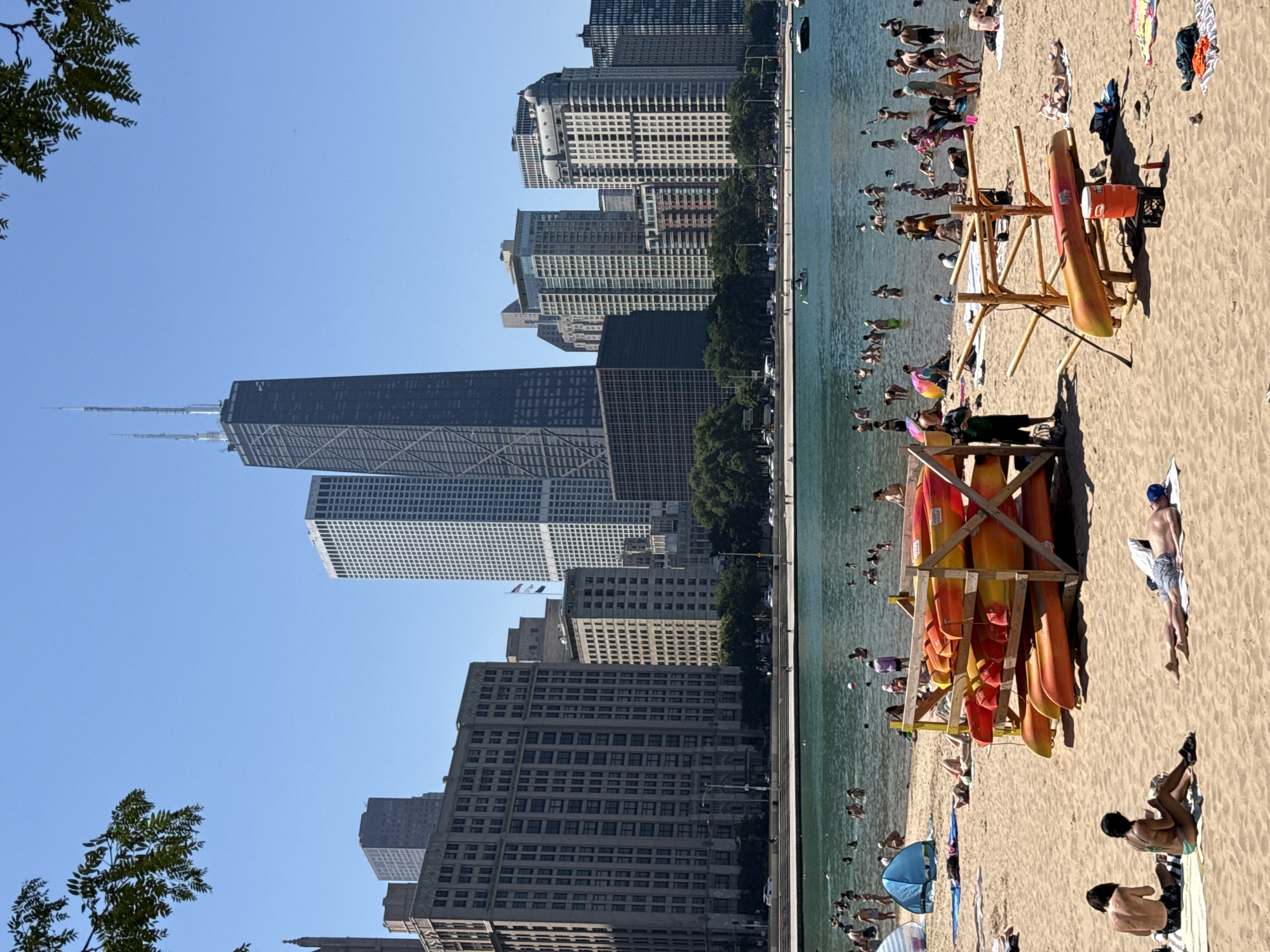 This image captures a vibrant summer scene at North Avenue Beach in Chicago, Illinois. The iconic John Hancock Center, a prominent skyscraper in the city's skyline, serves as a backdrop to the bustling beach activity. In the foreground, a sandy beach is dotted with people enjoying the sun and water. Several colorful kayaks are neatly stacked on wooden racks, indicating recreational opportunities. The clear blue water of Lake Michigan is filled with swimmers and bathers, some wading near the shore while others are further out. Beachgoers are seen lounging on towels, sunbathing, and socializing. The overall atmosphere is one of leisure and summer enjoyment.