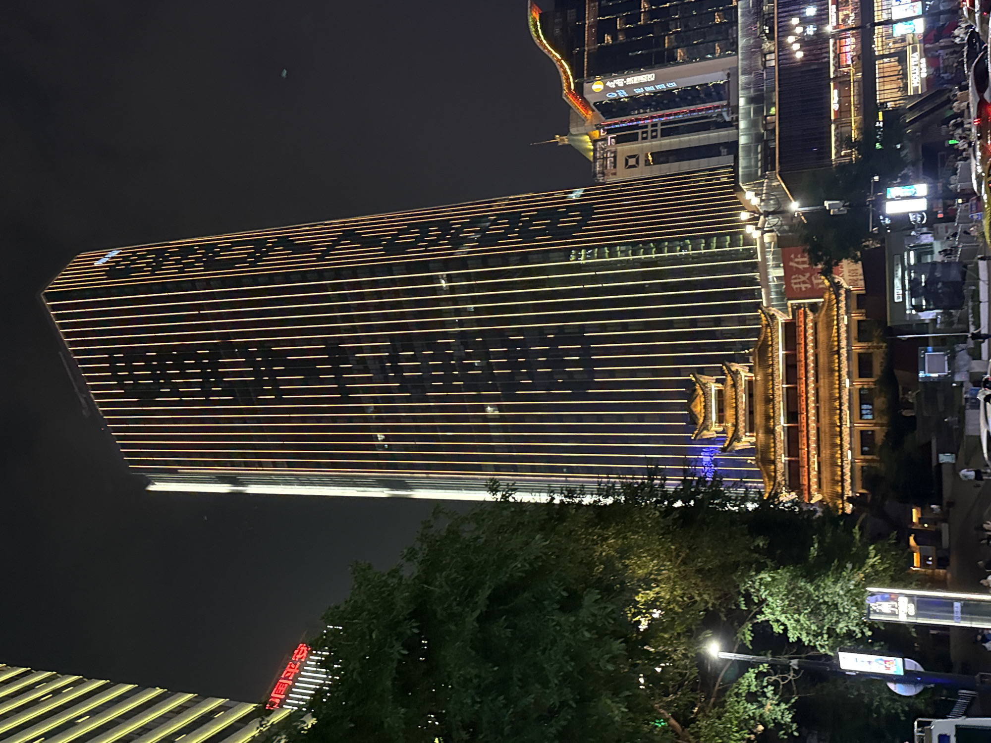 A low-angle, nighttime photograph captures a vibrant urban scene in Yuzhong District, China. Dominating the frame is a tall, modern skyscraper adorned with vertical, illuminated lines of warm light against its dark facade. To its left, another building features horizontal yellow lights. In the foreground, a traditional Chinese-style building with multiple tiered roofs, also illuminated, stands out. Trees with lush green foliage are visible in the mid-ground, framing the architectural elements. Streetlights cast a glow on the scene, and the lower portion of the image shows a street with parked cars and a few pedestrians. The sky is dark and appears clear. A sign with red Chinese characters is visible on the traditional building.