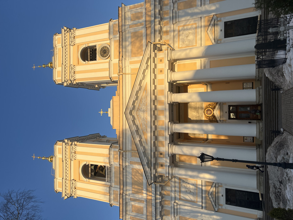 The image displays the facade of the Prince Vladimir Cathedral (Knyaz-Vladimirsky Sobor) in Saint Petersburg, Russia, under a clear, bright blue sky with low-angle sunlight. The large, classical-style building is predominantly light yellow with white architectural details. It features two prominent bell towers, one on each side, topped with golden onion domes and golden Orthodox crosses. The left tower contains multiple bells, while the right tower displays a visible clock face. A central structure above the main portico also bears a golden Orthodox cross.
The main facade includes a large portico with four tall, white, fluted Corinthian columns supporting a classical pediment adorned with relief sculpture. Rectangular panels with additional relief sculptures are present on the building's walls between the columns and the side doors. The central main entrance consists of a double wooden door, above which an ornate golden emblem is situated. Two darker, arched side doors, likely metal, are located on either side of the portico.
One individual, dressed in dark clothing, walks from right to left on the stone steps leading to the left side door. A second figure is partially visible within the central doorway. In the foreground, slightly left of center, a black, ornate street lamp post with a lantern is visible. Patches of melting snow and slush cover the ground, particularly at the base of the building and along the paved pathway leading to the main entrance, indicating a winter or early spring season. To the lower right, a section of a black wrought-iron fence encloses what appears to be a grave or memorial with a dark headstone and a white Orthodox cross. Bare tree branches are visible in the upper left corner of the frame. The strong sunlight casts prominent shadows on the architectural features.