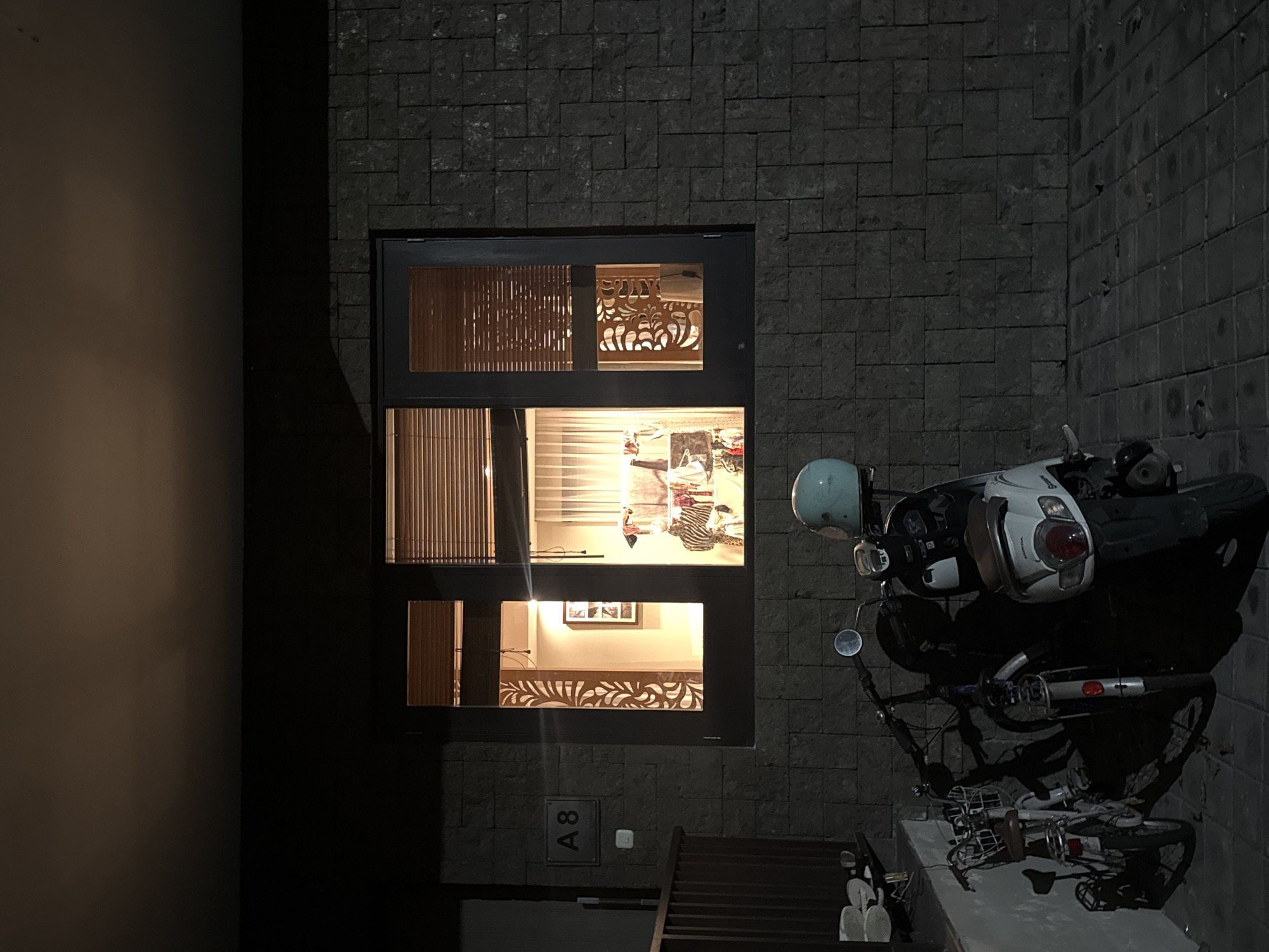 A nighttime exterior shot shows the corner of a building with a wall made of gray stone blocks and a large dark window. The window has three sections, with the top and bottom sections containing wooden slat blinds and decorative carved wooden panels. The middle section of the window allows a view into a brightly lit room with a lamp, furniture, and clothing. A light-colored scooter with a pale blue helmet rests against the stone wall, and a bicycle is parked nearby. A small plaque with the number "A8" is visible on the wall beneath the window. A bench sits in front of the wall below the motorcycle.