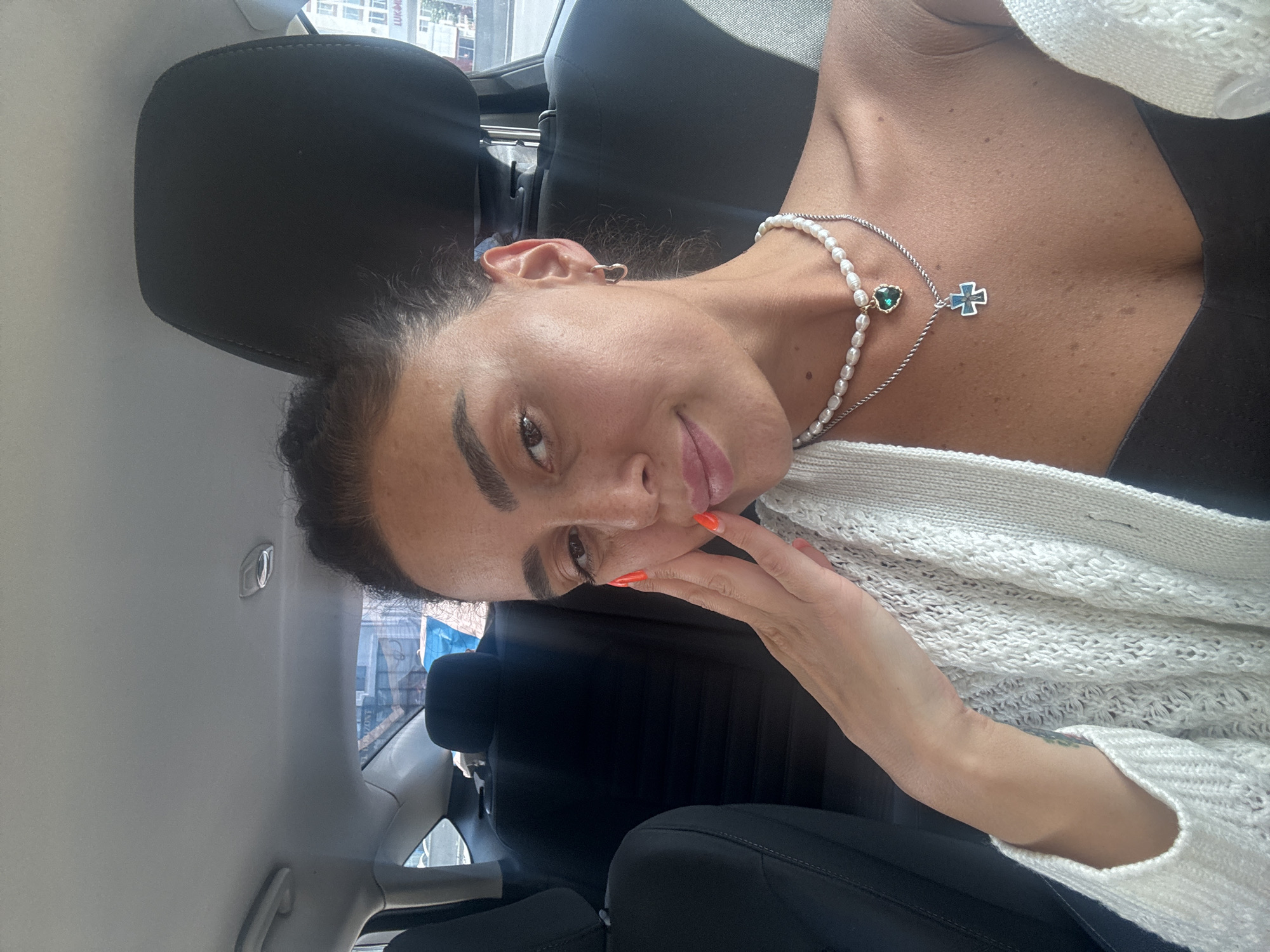 A woman is sitting in the driver's seat of a car. She is wearing a white knit cardigan over a black top. She has on a pearl necklace with a green heart-shaped pendant and a silver chain with a blue cross pendant. Her fingernails are painted orange. She is resting her right hand on her cheek and looking at the camera. The interior of the car is visible, including the headrests and part of the dashboard. Outside the car, buildings and a street are visible. The image was taken in Tbilisi, Georgia.