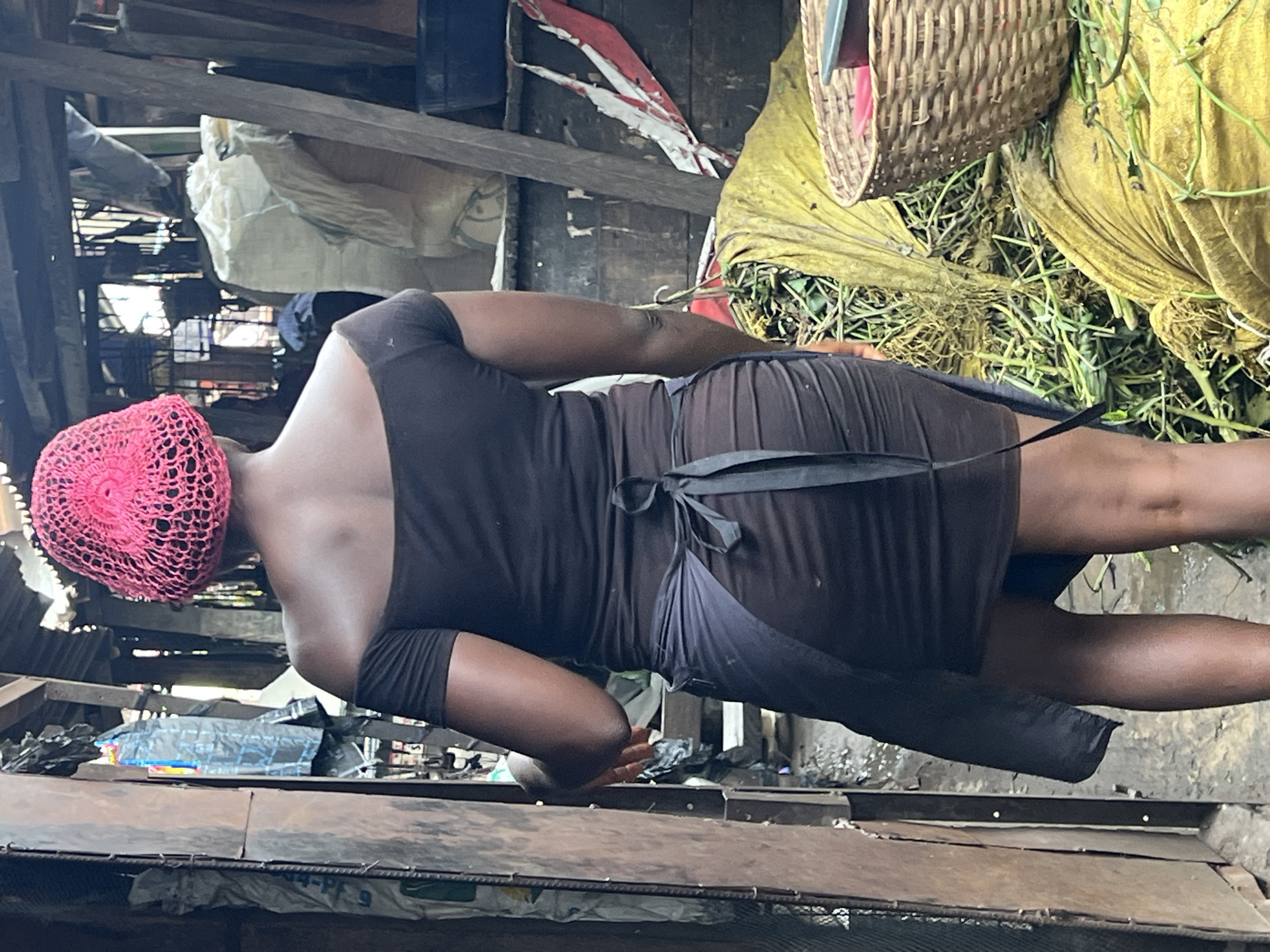 The image depicts a market scene in Nkpor, Nigeria, focusing on a Black woman from behind. She wears a bright pink netted head covering and a dark, off-the-shoulder dress with ruching around the skirt. A dark gray apron is tied around her waist with a bow. She is positioned in what appears to be a narrow aisle between market stalls, with wooden structures and various items visible in the background. To her right, large bundles of yellow fabric are piled up, one of which contains green vegetation, and a woven basket sits on top of it. The ground is damp and appears to be a dirt or concrete surface. The overall impression is that of a busy, functional market environment.
