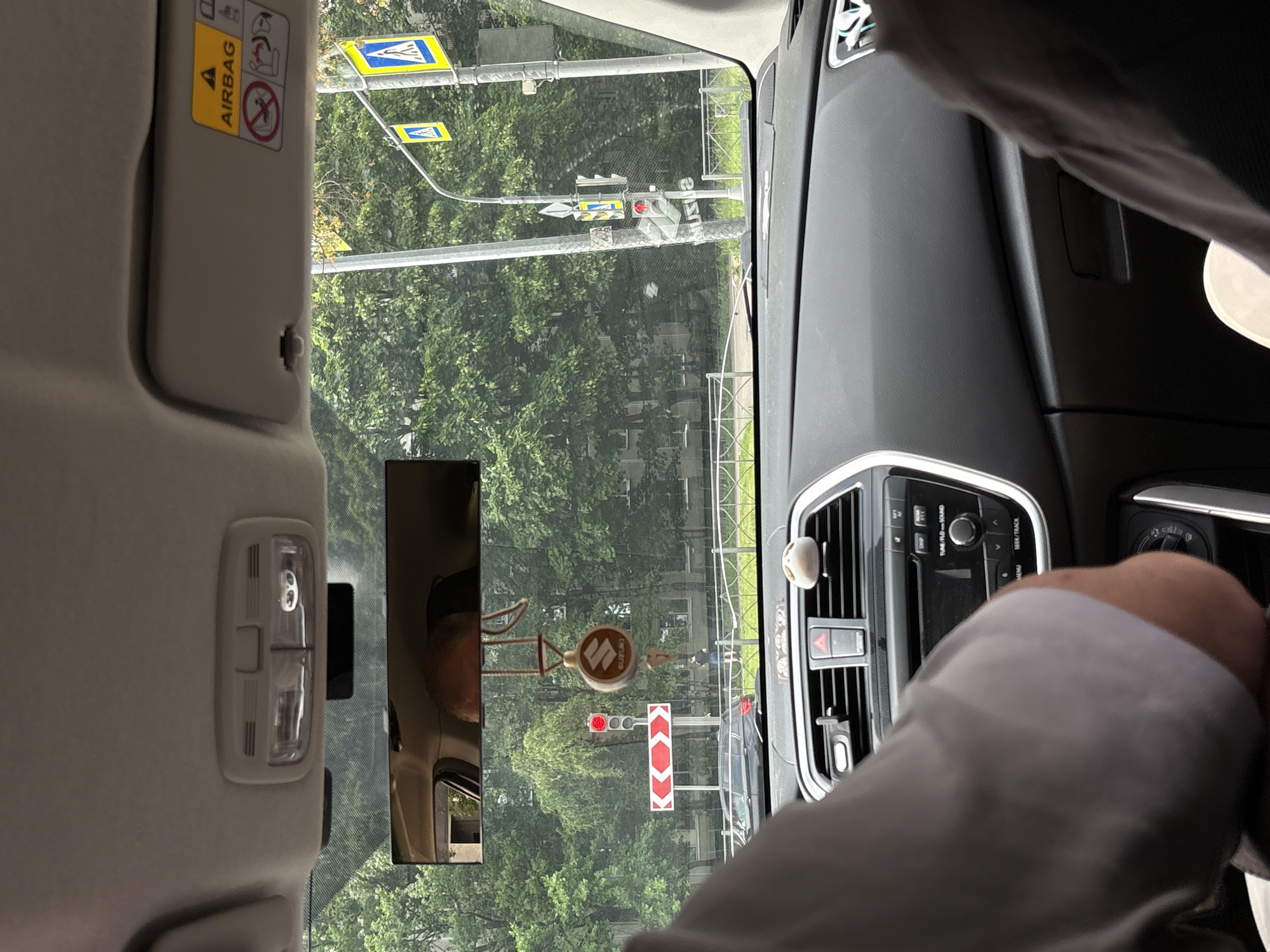 This image, taken from inside a car in Saint Petersburg, Russia, captures a view through the windshield and rearview mirror. The scene is dominated by lush green trees lining a street, with buildings visible in the background. A traffic light shows red, indicating a stop. To the left of the traffic light is a red and white directional sign with double arrows pointing left.
The car's interior features a rearview mirror reflecting the head of a person and a dashboard with a Suzuki-branded air freshener hanging from the mirror. The dashboard also displays a car stereo system and air vents. A yellow "AIRBAG" warning sticker is visible on the sun visor. Two traffic lights and pedestrian crossing signs are visible outside the car. A person in dark clothing is seen standing on the sidewalk in the distance.