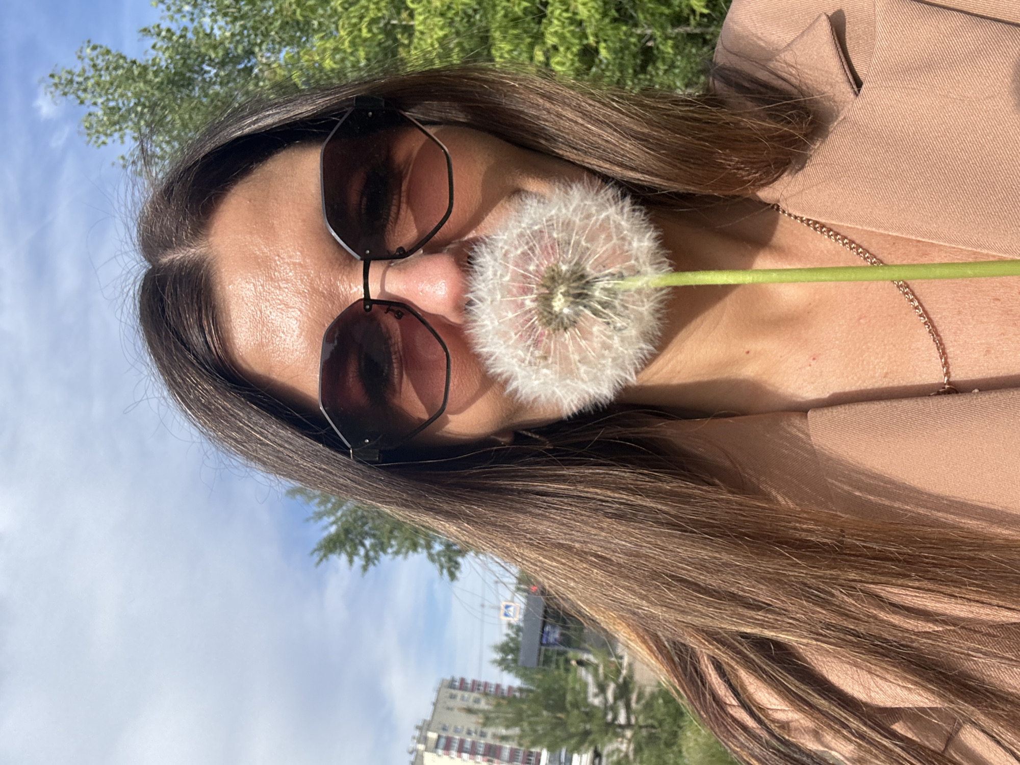 The image presents a close-up, vertically oriented shot of a person with long, flowing dark brown hair. They are wearing dark, octagonal-framed sunglasses and a light beige or tan blazer. A fully mature white dandelion puffball, with its delicate seeds ready to disperse, is held against their face, specifically covering the lower part of their nose and mouth. The dandelion's green stem extends horizontally to the right. A thin gold chain necklace is visible on the person's chest.
The setting is outdoors under a bright blue sky with scattered white clouds, indicating a clear, sunny daytime. Green tree foliage is visible in the background, along with blurred, distant multi-story residential buildings, suggesting an urban or suburban park or street environment in Chaikovsky, Russia. The light on the person and their attire suggests bright sunlight. There is no visible text in the image.