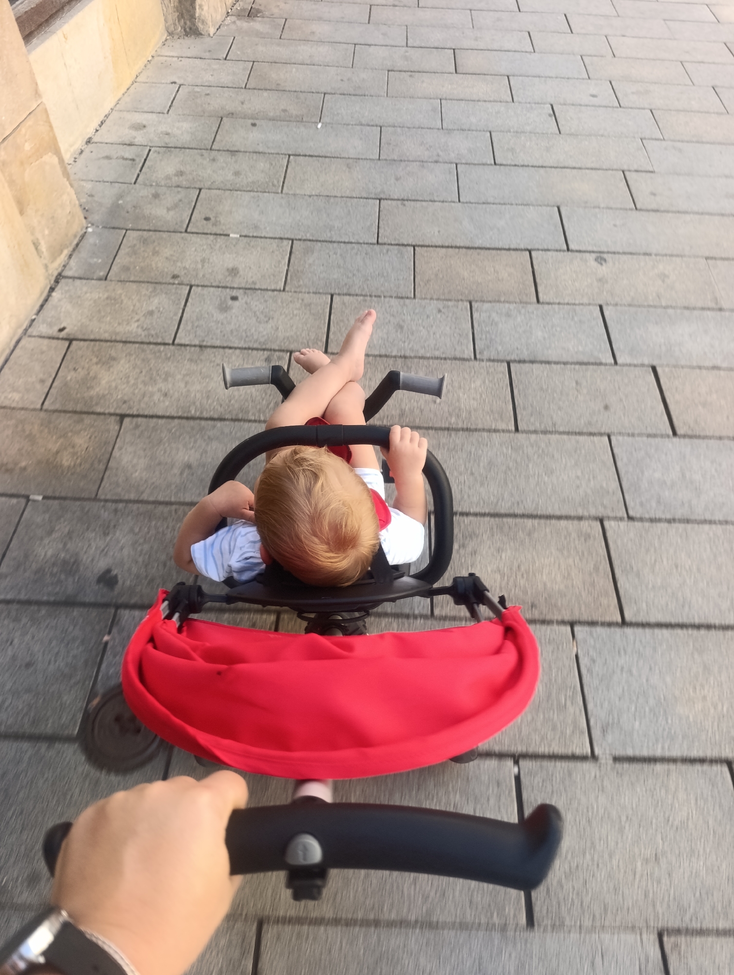 A high-angle, full shot shows a person's hand holding the handlebar of a tricycle, with a young child seated in the tricycle, their legs crossed at the ankles. The child has fair hair and is wearing a striped shirt with a red collar. A red canopy is attached to the tricycle. The tricycle is on a paved walkway made of rectangular gray stones. The background is a blur of these gray stones and a beige wall on the left. The scene appears to be outdoors, possibly in a city, given the paved walkway and wall.