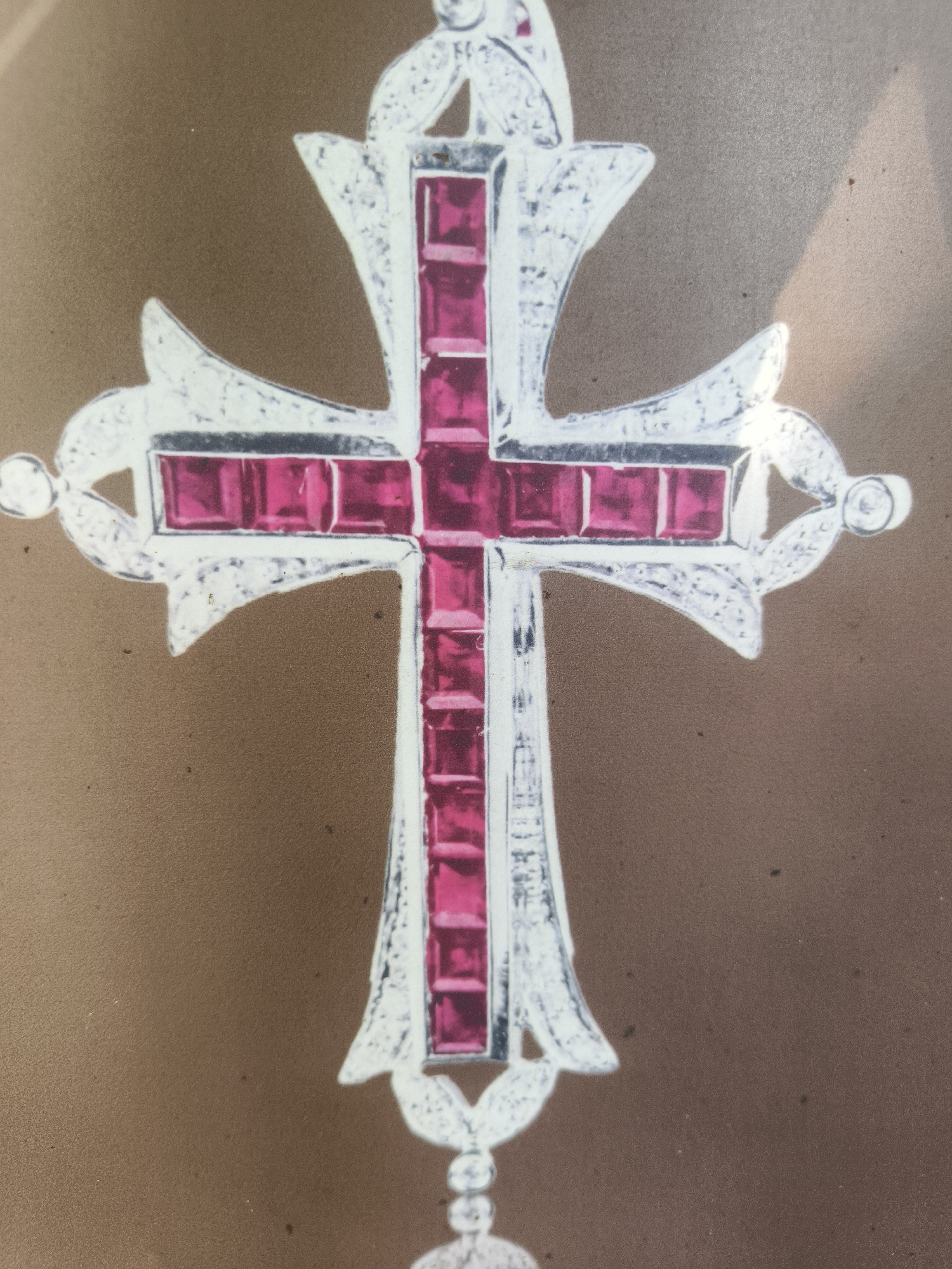 A close-up, overhead shot shows a detailed illustration of a cross pendant. The cross is primarily composed of a silver-colored metal, intricately designed with what appear to be small, square pink gemstones embedded within its structure. The gemstones line the arms and shaft of the cross, creating a vibrant, repeating pattern. The metalwork features decorative flourishes and ornate detailing, suggesting a piece of fine jewelry. The background is a solid, mottled brown color, likely representing paper or a similar flat surface on which the illustration is presented. There is a subtle shadow cast on the right side of the background, indicating an external light source. The image focuses solely on the pendant, with no other objects or subjects visible. The location is Kuta Selatan, Indonesia.