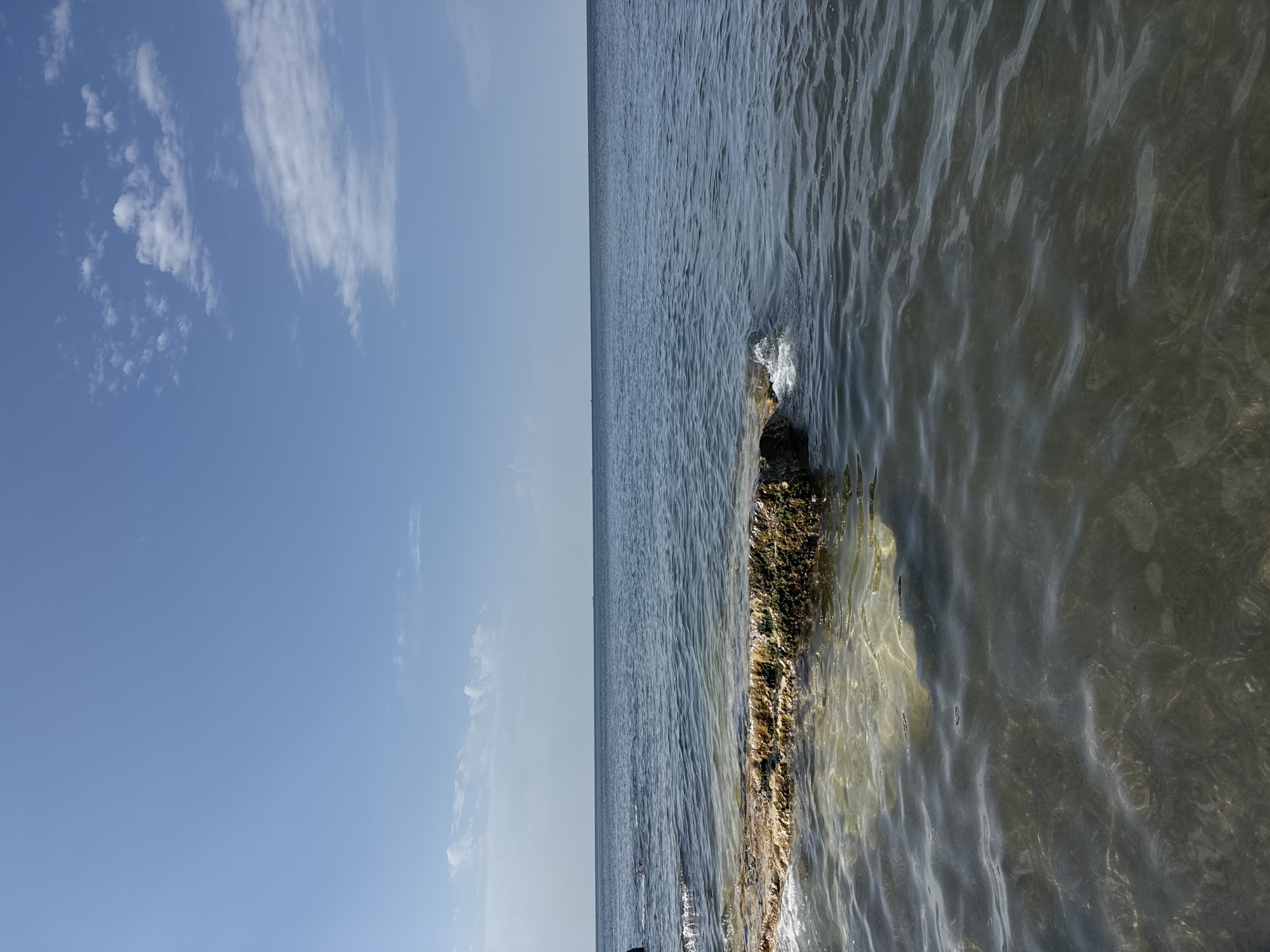 This image depicts a serene seascape with a clear blue sky dotted with wispy white clouds. The ocean stretches to the horizon, its surface rippling gently. In the foreground, a partially submerged rock formation, covered in patches of algae and moss, breaks the water's surface. Waves are seen lapping around the rock. The water appears clear, allowing a glimpse of the rocky seabed below. This scene is characteristic of the coastline along Aktau, Kazakhstan, a city known for its Caspian Sea shore. No people are present in the image.