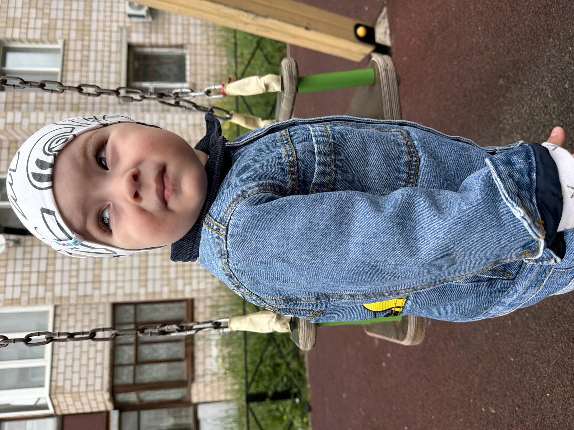 The image shows a young child wearing a denim jacket and a patterned beanie, standing on a playground in Votkinsk, Russia. The child is looking upwards and to the right. The playground features a swing set with metal chains and wooden supports, and the ground is covered in a reddish-brown material, likely rubberized safety surfacing. In the background, a light-colored brick building with multiple windows is visible. The overall setting appears to be an outdoor urban or suburban environment.