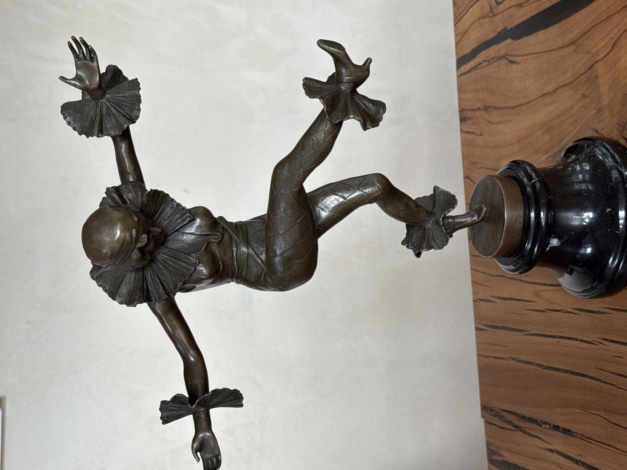 A bronze sculpture of a jester or acrobat is mounted on a stand. The figure is in mid-air, with one leg extended upward and attached to the stand. The figure is wearing a ruffled collar and wrist cuffs and diamond patterned leggings. The stand is made of black and gold/bronze metal. The background consists of a white wall and a wood panel.
There is no information in the image itself or the hints provided to indicate this image was taken in Steklyannyi, Russia.