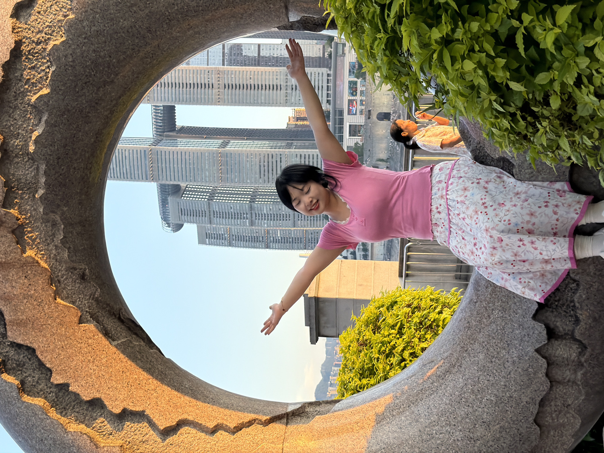 A young Asian woman is captured in a photograph, framed by a circular stone archway with a jagged, textured edge illuminated by warm, golden light. She is seated on a textured stone surface, wearing a bright pink short-sleeved shirt and a long, flowing skirt adorned with a delicate floral pattern in pink and blue. Her arms are outstretched upwards and outwards, and her eyes are closed, conveying a sense of joy and freedom.
Behind her, the urban landscape of Nan'an District, China, unfolds under a clear blue sky. A cluster of imposing skyscrapers dominates the background, with one particularly striking building featuring a skybridge connecting two towers. The architecture suggests a modern, bustling city.
To the right of the main subject, another woman is visible, seated further back and partially obscured by lush green foliage. She is wearing a light-colored, patterned shirt and appears to be looking down, perhaps at something in her hand.
The foreground is occupied by the textured stone arch and vibrant green bushes, creating a naturalistic frame for the urban vista and the people within it. The sunlight highlights the textures of the stone and the leaves, adding depth and warmth to the scene. The overall impression is one of serenity and engagement with the urban environment.