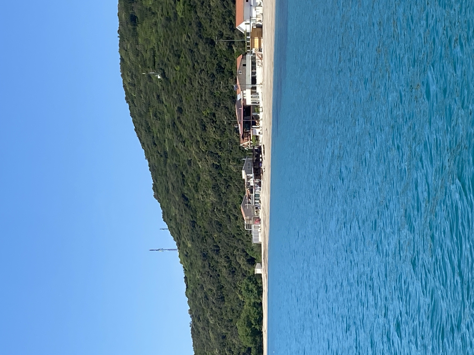 The image displays a serene coastal or lakeside landscape under a clear, bright blue sky. On the left, a steep, verdant hillside, densely covered with lush green trees, rises prominently. Towards the upper portion of the hill, two slender, pole-like structures, possibly communication towers or utility poles, are faintly visible against the sky.
At the base of the hillside, a narrow strip of light-colored sand forms a beach, creating a natural division between the vibrant green foliage and the tranquil water. Along this sandy expanse, a cluster of low-rise buildings is situated. These structures, likely houses or small accommodations, exhibit varied architectural styles, some with white facades and others with warmer, earthy tones. Several utility poles with overhead lines are interspersed among the buildings, indicating established infrastructure.
To the right of the beach and buildings, a vast expanse of clear, turquoise-blue water extends across the frame. The water surface shows gentle ripples, characteristic of calm sea or lake conditions, and its transparency suggests a clean environment.
The scene depicts a bright, sunny day, likely during morning or early afternoon hours, given the absence of clouds and strong illumination. The overall atmosphere conveys peace and natural beauty. No people are visible, and no specific activity or event is taking place. The location, Prijevor, Montenegro, suggests this is a view of the Adriatic coast or a large body of water in the region. There is no visible text in the image.