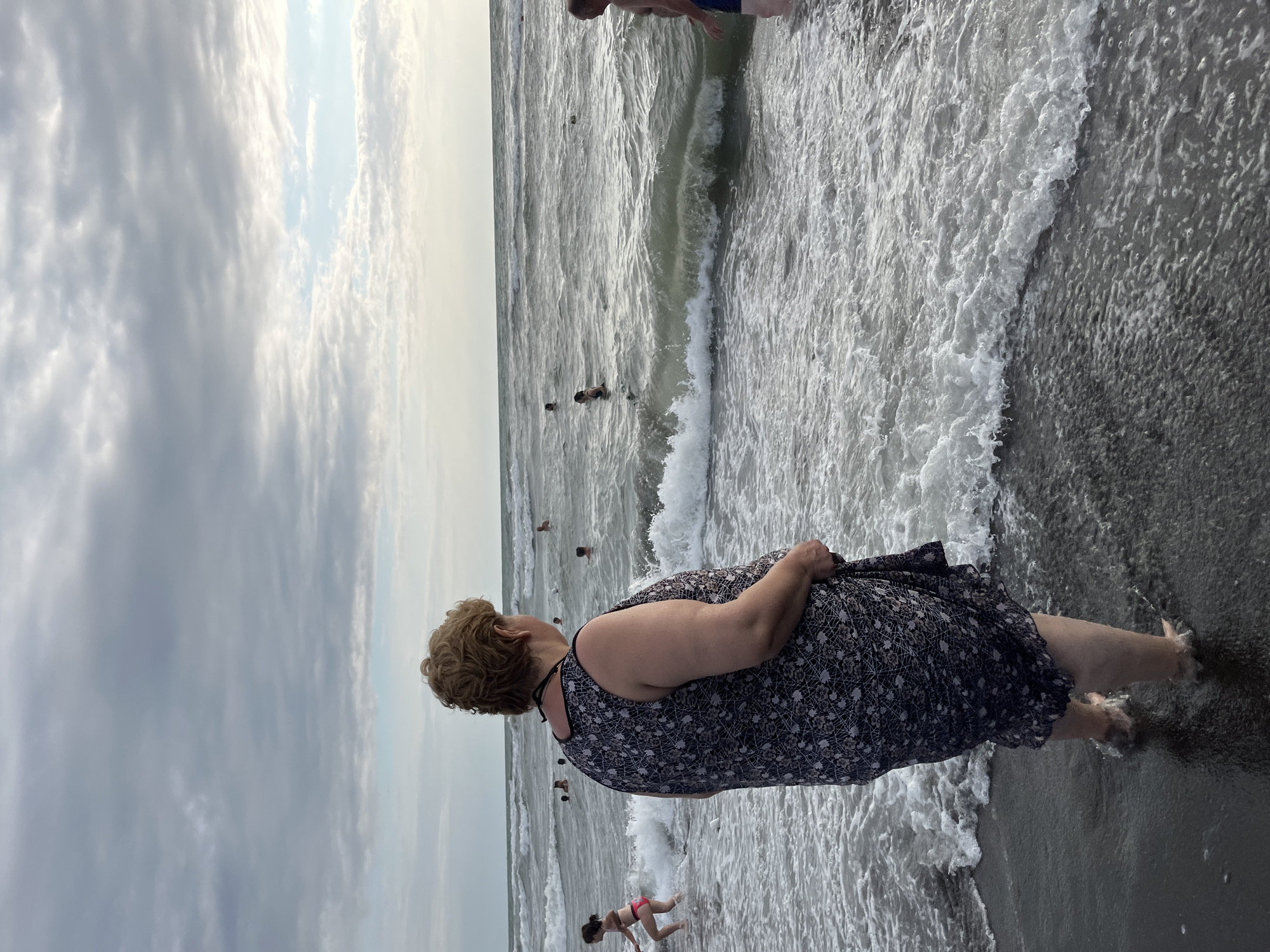 An overcast day at Ureki, Georgia's magnet sand beach. A woman stands in the shallow water, facing away from the camera, her back to the viewer, dressed in a dark, floral-patterned dress. The sea is rough with moderate waves, and several people are visible in the water further out, swimming and playing. The sky is filled with scattered clouds.