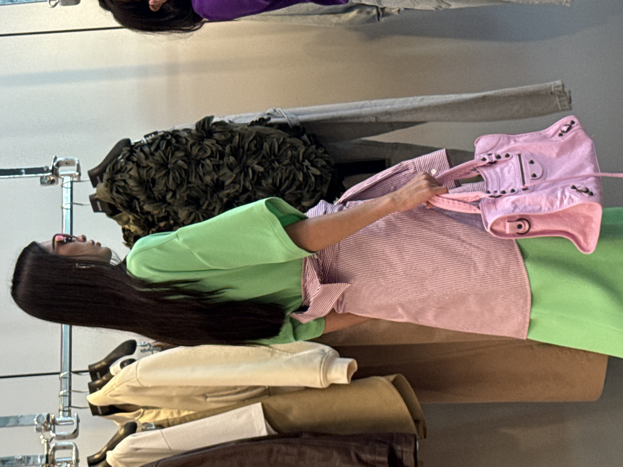 A woman with long dark hair, wearing a neon green shirt and a pink and white striped skirt, stands in a retail environment, likely a clothing store. She is holding a bright pink handbag. Behind her, clothes are displayed on a rack. The scene appears to be indoors with a light-colored wall in the background. The woman is facing to the right, partially obscuring her face with her hair. The lighting is bright and even, suggesting an indoor retail setting.