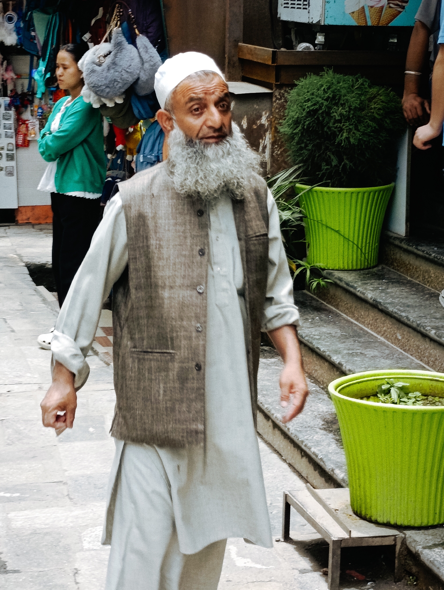 An elderly man with a white skullcap and a long white beard, wearing a grey vest over a light-colored kurta, walks down a cobblestone path in Manali, India. He has his right hand slightly outstretched. Behind him, a young woman in a green jacket and black pants stands with her arms crossed, facing left. The background shows a market stall with various items hanging, including stuffed animals and clothing. A large, bushy green plant in a bright green pot is visible to the right, as are stone steps and another green pot with smaller plants. The overall scene depicts a street in a bustling market area.