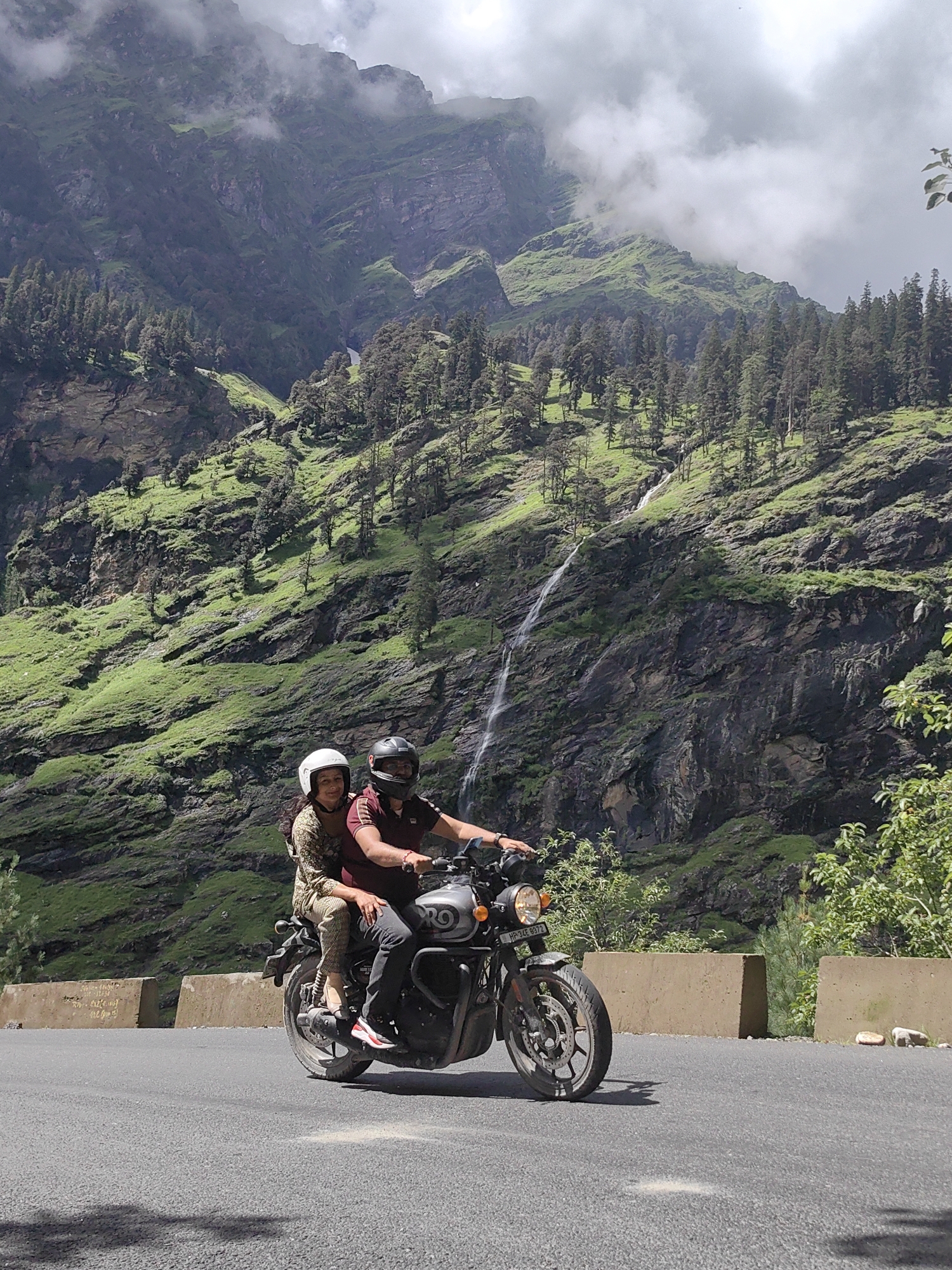Two people, a man and a woman, are riding on a motorcycle on a paved road. The man is driving, and the woman is riding as a passenger behind him. Both are wearing helmets. They are positioned in the lower half of the frame, with the road occupying the bottom portion and a steep, green, mountainous landscape filling the background.
The mountain slopes are covered in lush green grass and dotted with numerous coniferous trees. A thin waterfall cascades down the rocky face of the mountain behind the motorcycle. The sky is partially cloudy, with patches of white clouds obscuring the very top of the mountains. The overall impression is of a scenic mountain road, possibly in a rural or remote area. The time of day appears to be daytime, with moderate lighting.
The motorcycle is a dark color with a silver fuel tank and silver wheels. The license plate is visible and reads "HP-34-CF-9972". The people are dressed casually. The woman is wearing a patterned dress and pants, while the man is wearing a maroon t-shirt and dark pants. Their expressions are neutral. The setting appears to be Burwa, India, based on the provided location context.