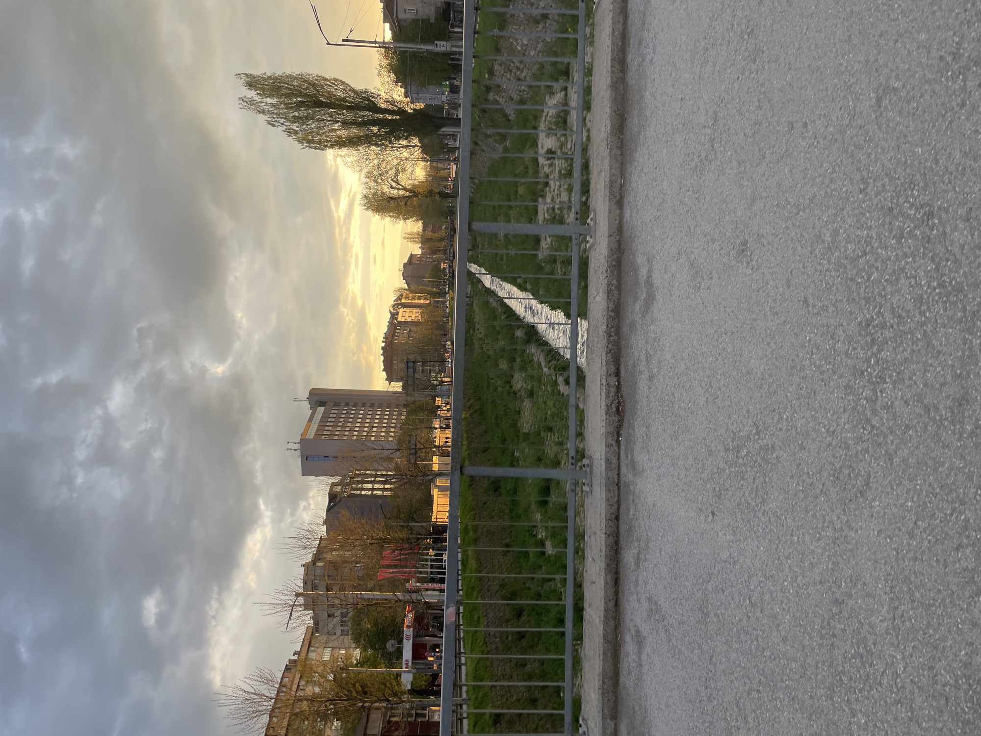 The image presents an urban landscape in Sofia, Bulgaria, captured during the golden hour, likely sunrise or sunset. The scene is viewed from a paved surface, possibly a sidewalk or road, which occupies the right side of the frame and shows a granular texture.
A dark grey metal fence with vertical bars runs horizontally across the midground, separating the paved area from a strip of green land. Beyond this fence, a narrow, light-colored channel, appearing as a dry streambed or shallow canal, cuts through a grassy area. Trees are scattered throughout the mid-ground and background; some are bare, suggesting an early spring or late autumn setting, while others have green foliage.
In the background, a series of buildings lines the horizon. A prominent, tall, modern building with many windows and a light-colored facade stands out, reflecting the warm, low sunlight. Other residential or commercial structures of varying heights and older styles are also visible, partially illuminated by the warm glow.
The sky above is largely covered by clouds, ranging from light grey to darker tones. A bright, golden light from the sun, positioned low on the horizon, breaks through the clouds, casting a warm hue across the scene and highlighting the edges of the clouds and buildings.
There are no people or specific activities depicted. The scene is static, capturing a moment of serene urban tranquility. No discernible text is visible within the image.