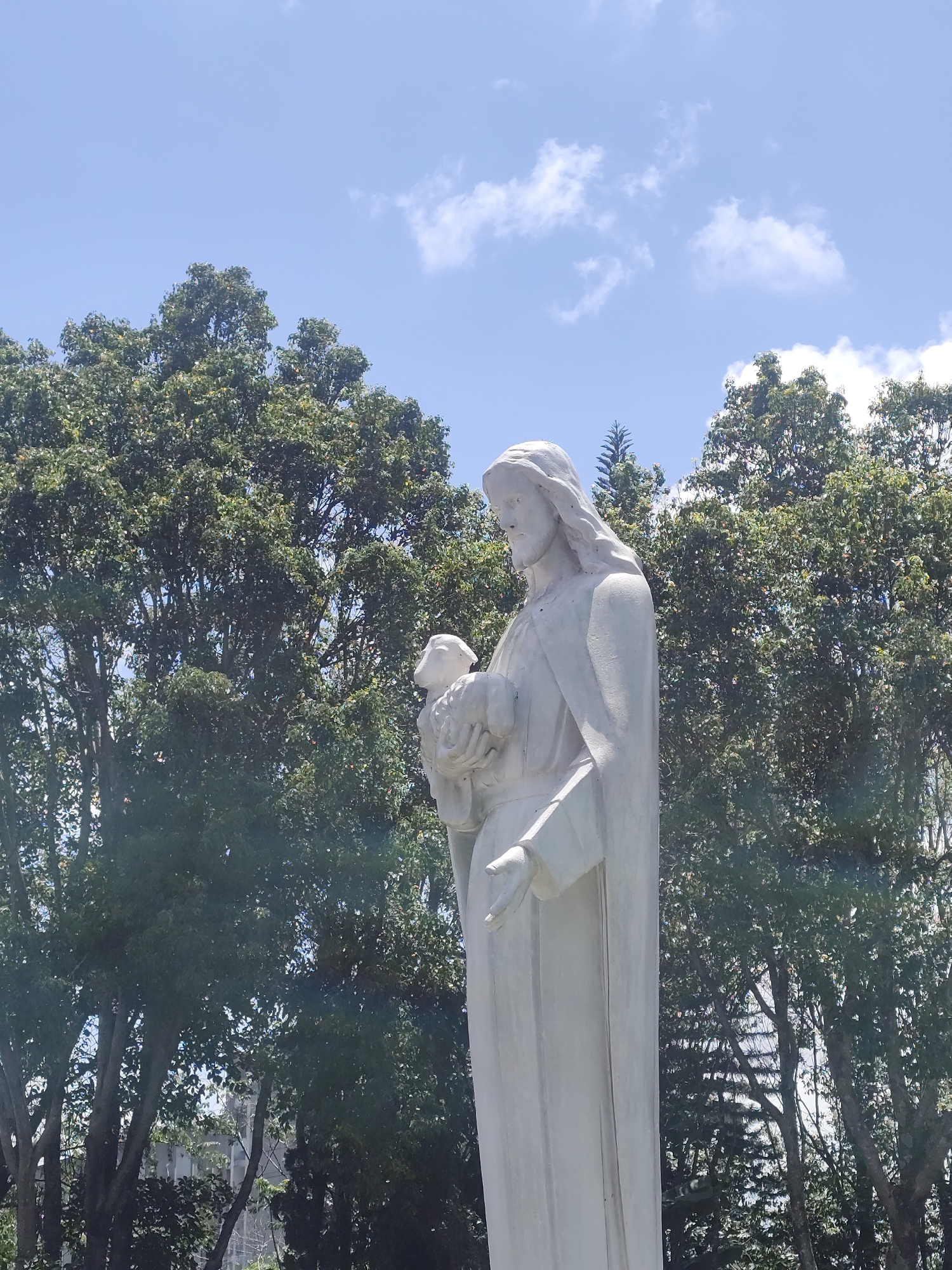 This image shows a white stone statue of Jesus Christ holding a lamb. The statue is positioned outdoors, with a clear blue sky and fluffy white clouds visible in the background. Lush green trees surround the statue, with some buildings peeking through the foliage in the distance. The lighting suggests it is daytime, with the sun casting a gentle light on the scene. This statue is located at the Dinh Thu Hoang De (Palace of the King) in Da Lat, Vietnam.