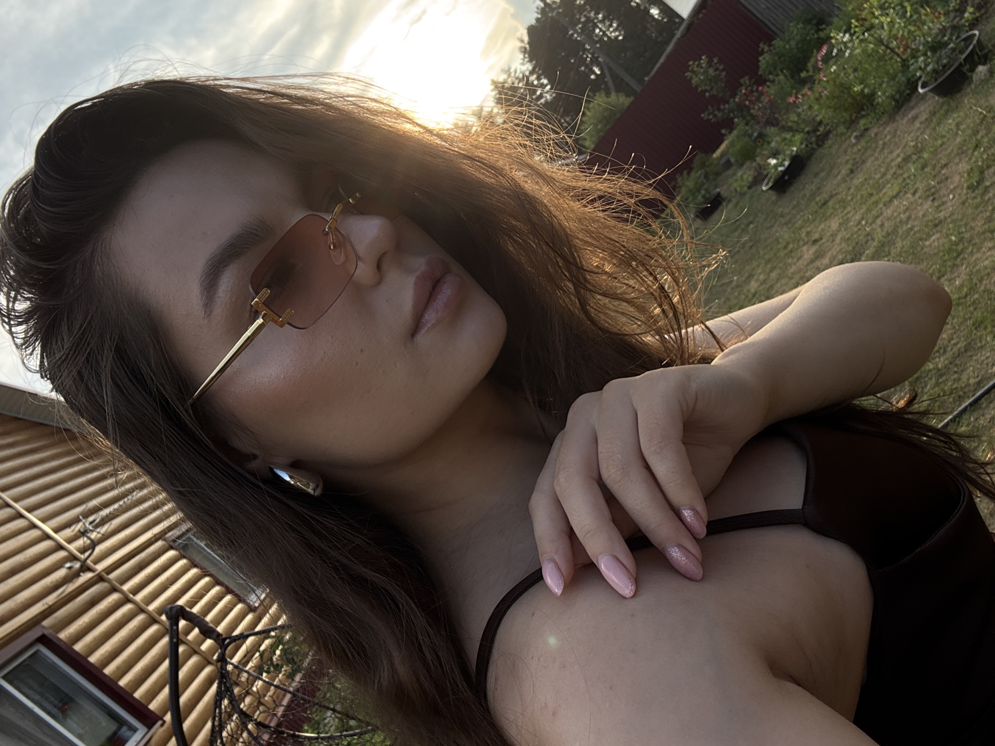 A young woman with long, dark hair and light skin is pictured outdoors in what appears to be a residential backyard in Rusalevka, Russia. She is wearing a dark brown camisole and large, gold-rimmed sunglasses with rose-tinted lenses. Her left hand, with light pink, glittery nail polish, is resting on her upper chest and shoulder. In the background, a wooden house with striped siding is visible, along with a garden area with plants in pots and a red fence. The lighting suggests it is late afternoon or early evening, with the sun casting a warm glow. The overall impression is a casual, sunlit moment.
