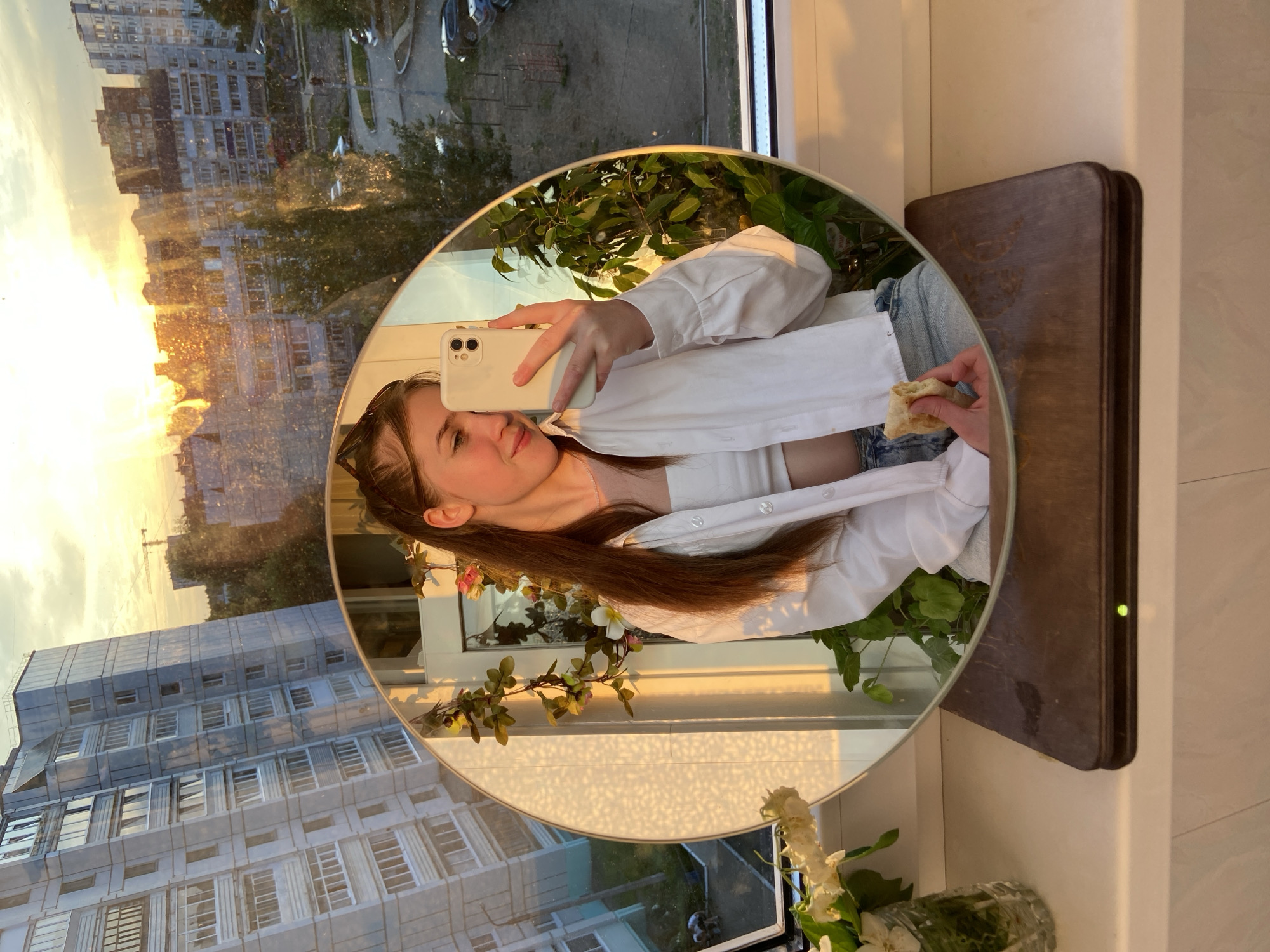A young woman with long brown hair, wearing a white crop top and an unbuttoned white shirt, takes a selfie in a round mirror. She is holding a piece of food in her right hand and a smartphone in her left. The mirror reflects her and a potted plant. In the background, through a window, a city skyline is visible at sunset, with apartment buildings, trees, and a construction crane. The scene appears to be set on a windowsill in Chaikovsky, Russia, suggested by the architectural style of the buildings and the landscape.