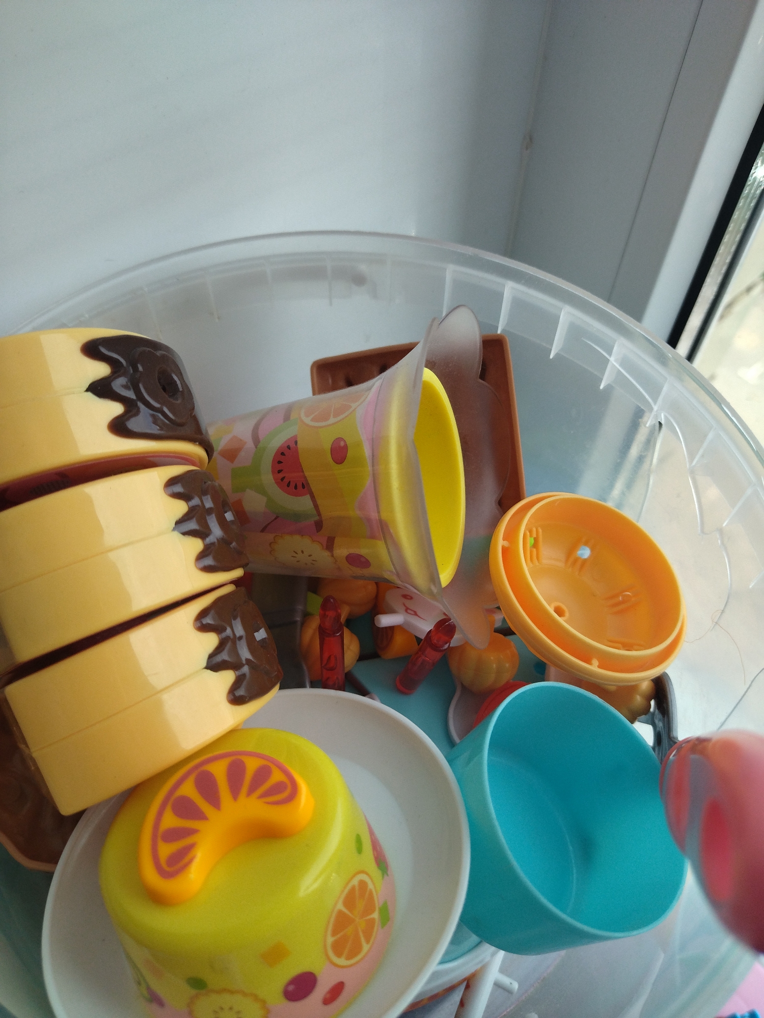 The image shows a collection of colorful plastic toys, likely for children, inside a clear plastic bin. Among the toys are what appear to be slices of a layered cake with chocolate frosting, a brightly colored cup or container with fruit designs, an orange bowl-like object, and a blue cup. Some smaller red and orange toy pieces are also visible. The toys are jumbled together within the bin, which is placed on a white surface, possibly a shelf or windowsill. The background is mostly white and out of focus, with a hint of light coming from what could be a window, suggesting an indoor setting. There is no visible indication of the city of Izhevsk, Russia, or any other specific location. The scene is static, with no active interactions or events taking place.