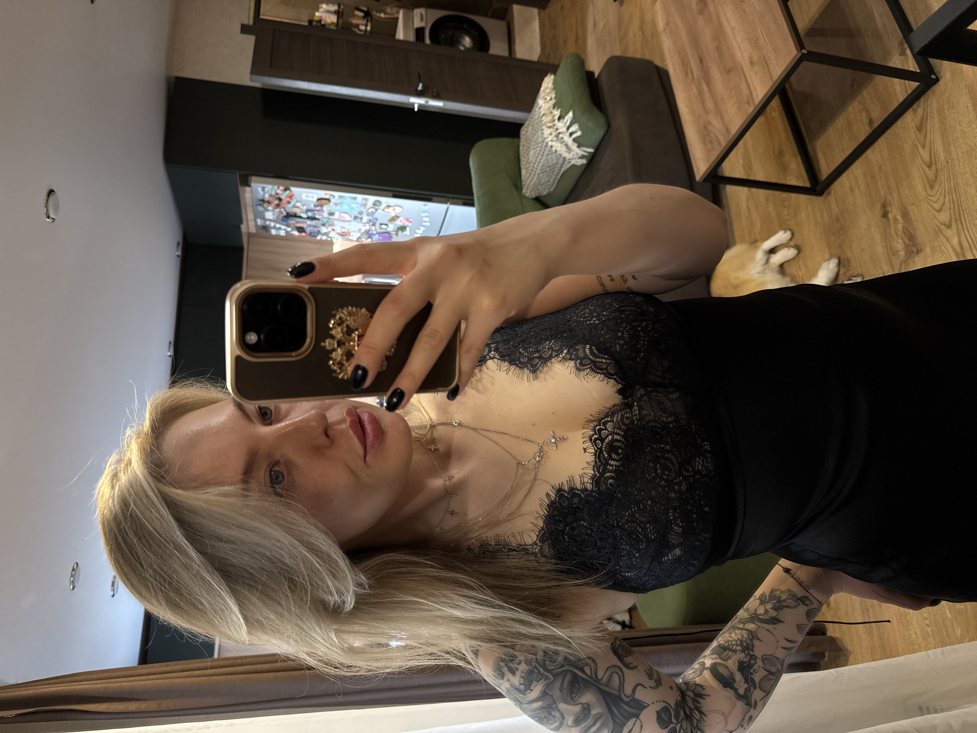 A woman with long blonde hair, wearing a black dress with lace detailing, is taking a selfie indoors. She holds a smartphone adorned with a decorative case. Her left arm features extensive tattoos. A small dog is visible near a couch in the background. The interior has a modern design, including a wooden table and a dark cabinet. The location is likely a private residence. No evidence confirms the city of Krasnoyarsk, Russia.