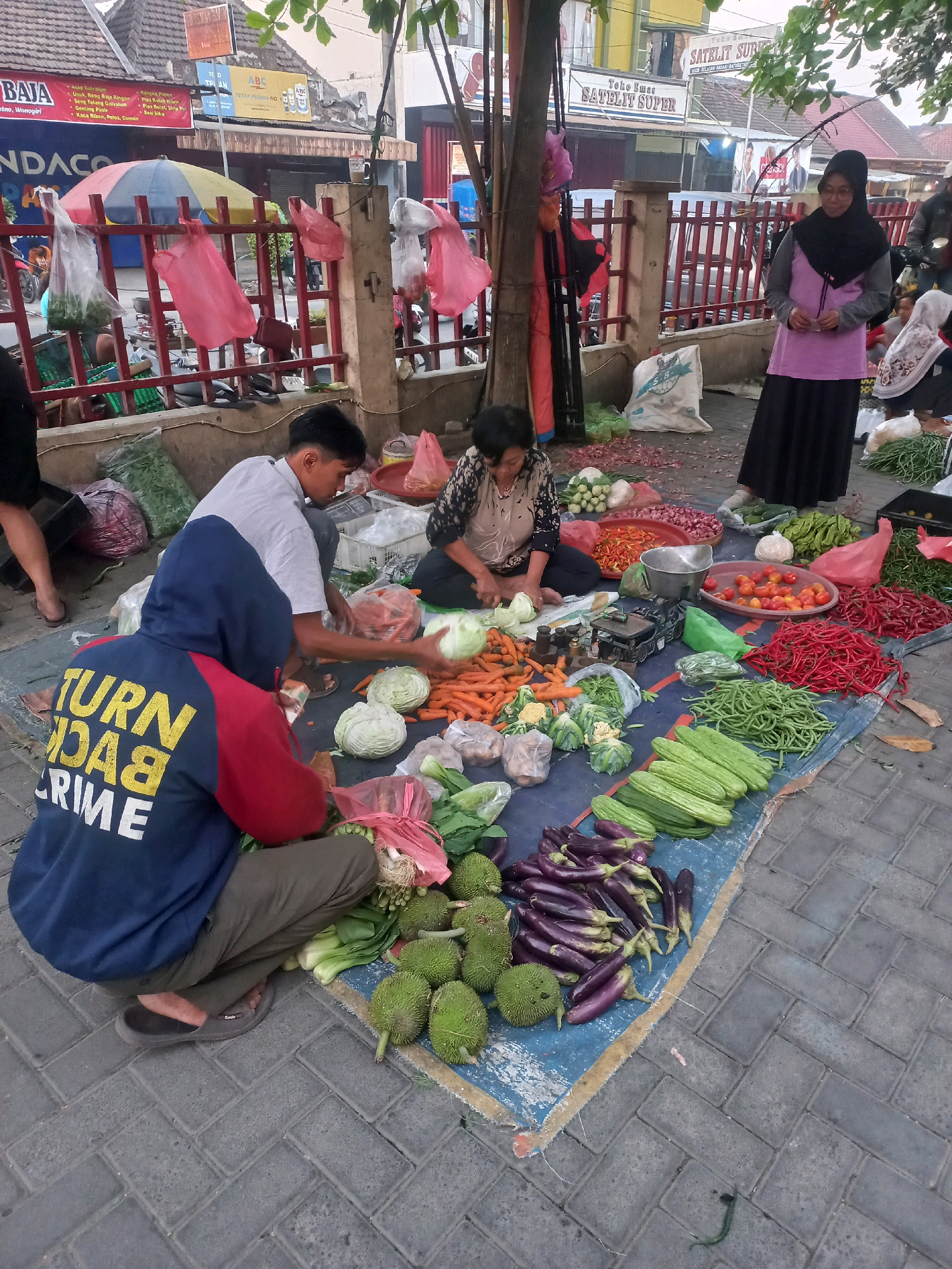 The image depicts an outdoor market scene in Baturetno, Indonesia. In the foreground, a young man wearing a blue hooded sweatshirt with yellow lettering that reads "TURN BACK CRIME" is crouching down, facing away from the camera. He is surrounded by a variety of fresh produce laid out on a blue tarp. Adjacent to him, another young man in a white shirt is sitting, seemingly sorting vegetables. A woman in a headscarf and purple top is standing to the right, and another woman in a dark headscarf and patterned top is seated and appears to be preparing produce.
The market stall displays an abundance of colorful vegetables, including carrots, cabbage, tomatoes, eggplant, cucumbers, green beans, and red chilies. Several plastic bags, some containing produce, are scattered around the area, along with larger sacks of goods. In the background, a red fence separates the market from a street with various shops and buildings. The overall impression is a bustling local market, characteristic of Indonesian street commerce.