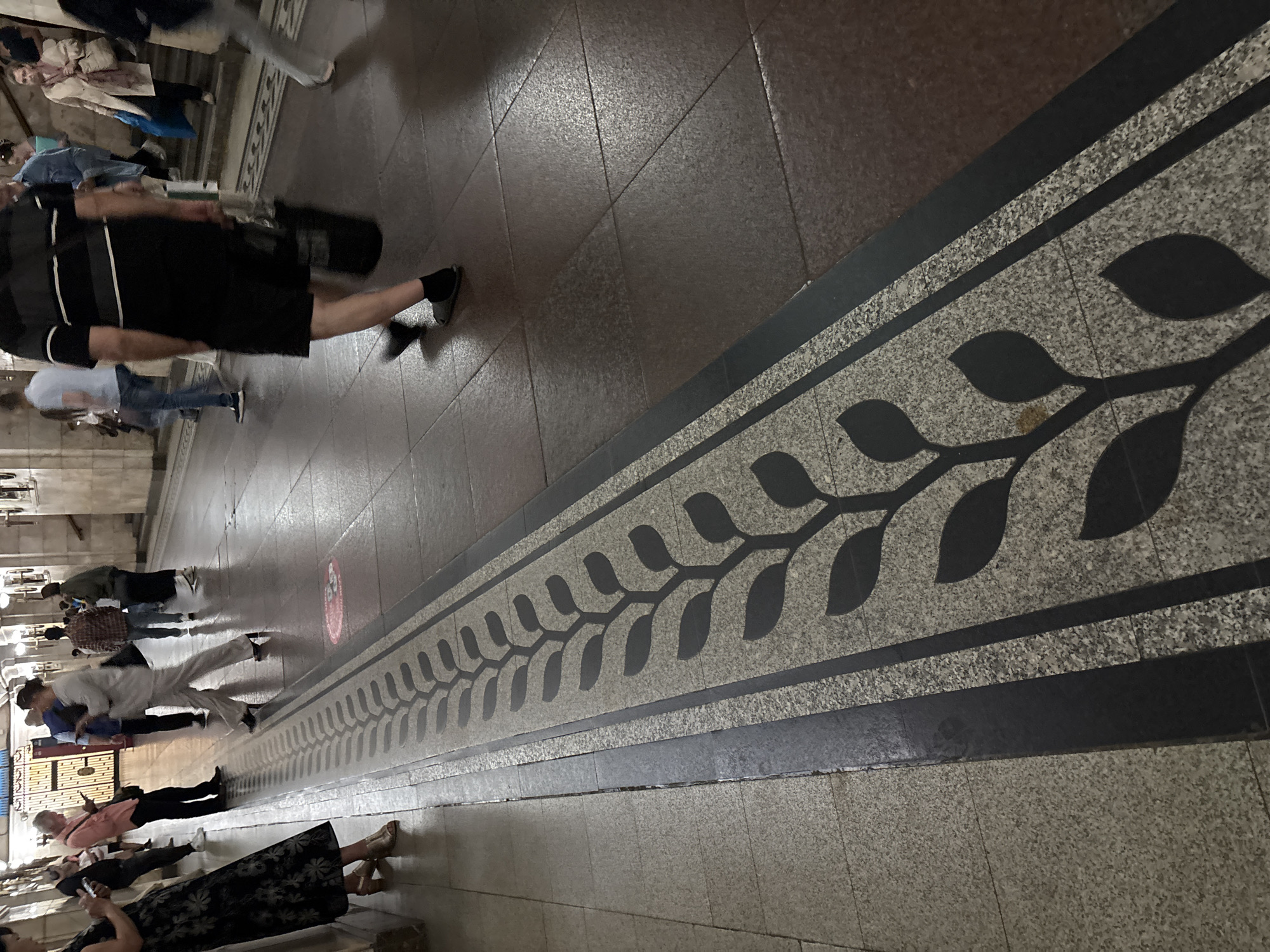The image captures a scene in a subway station, likely in Moscow, Russia, given the architectural style and the presence of decorative elements on the floor. The perspective is low, focusing on the floor with a distinct mosaic pattern of stylized leaves running along a pathway. Several people are visible in the background, some walking, others standing. One individual in the foreground, seen from the waist down, is wearing a black and white striped shirt and black shorts, walking with a black bag. The floor is tiled with a granite-like pattern, interspersed with the darker mosaic border. The lighting suggests an indoor environment, characteristic of a metro station. The overall impression is one of movement and daily transit within an urban setting.