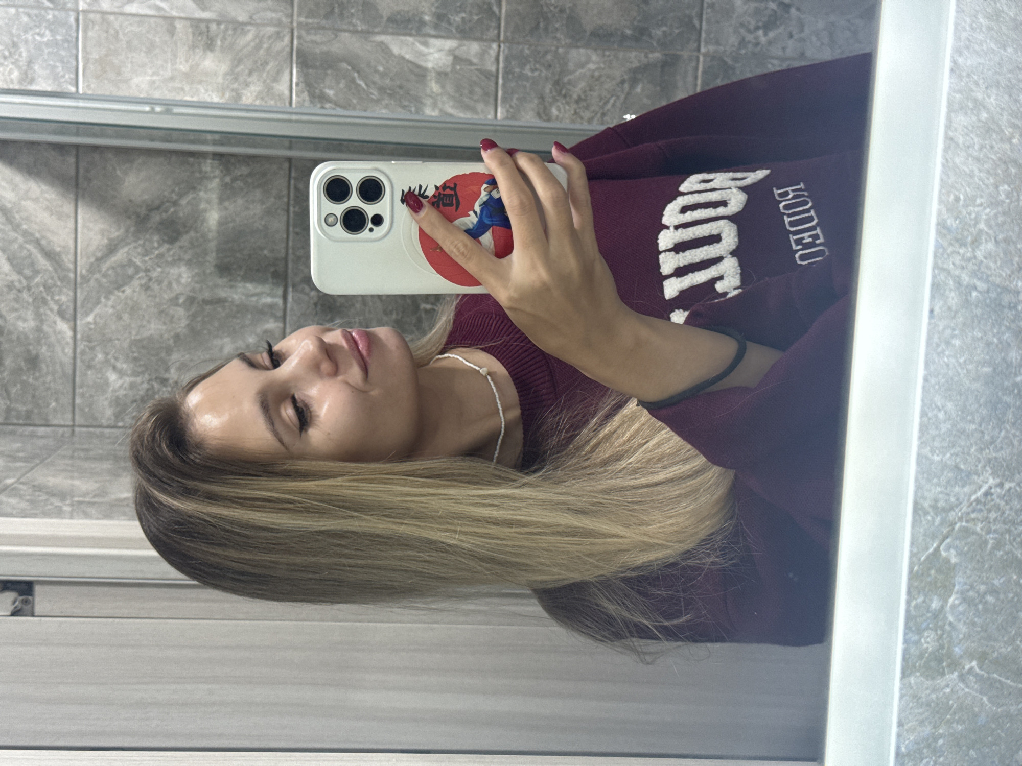 A young woman with long blonde hair takes a mirror selfie. She is wearing a maroon sweater with white embroidered text that appears to say "Rodeo" and "boom". Her phone, which has a white case with a red circular sticker featuring a person in a blue uniform, is held up to take the photo. She is wearing red nail polish and a simple white beaded necklace. The background shows grey tiled walls and a light-colored cabinet. The lighting suggests it is daytime. The woman has a slight smile. The location is Chaikovsky, Russia.