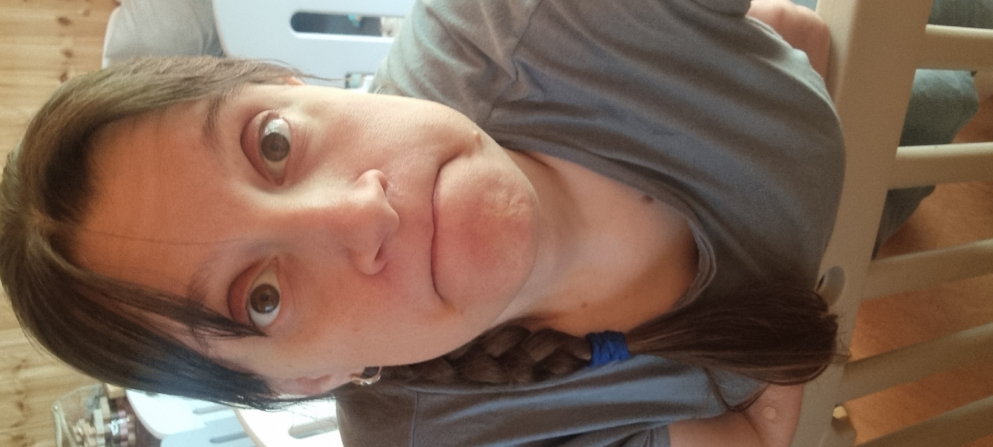 A woman with light skin, brown eyes, and brown hair with dark bangs in a braid with a blue hair tie is shown from a close, tilted perspective. She is wearing a gray t-shirt. The background includes wooden walls, white furniture, and wooden bars, possibly from a crib or cot. The location is not specified.